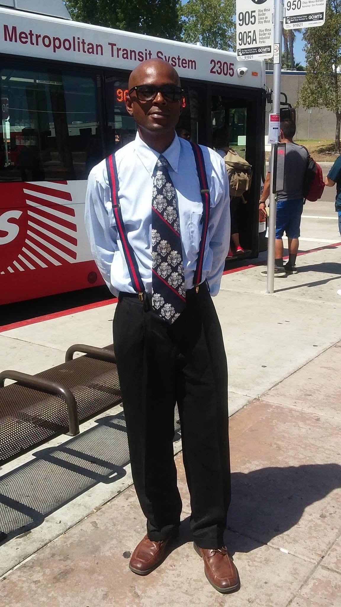 A man dressed in a formal shirt and tie stands at a bus stop during the day, waiting for a bus.