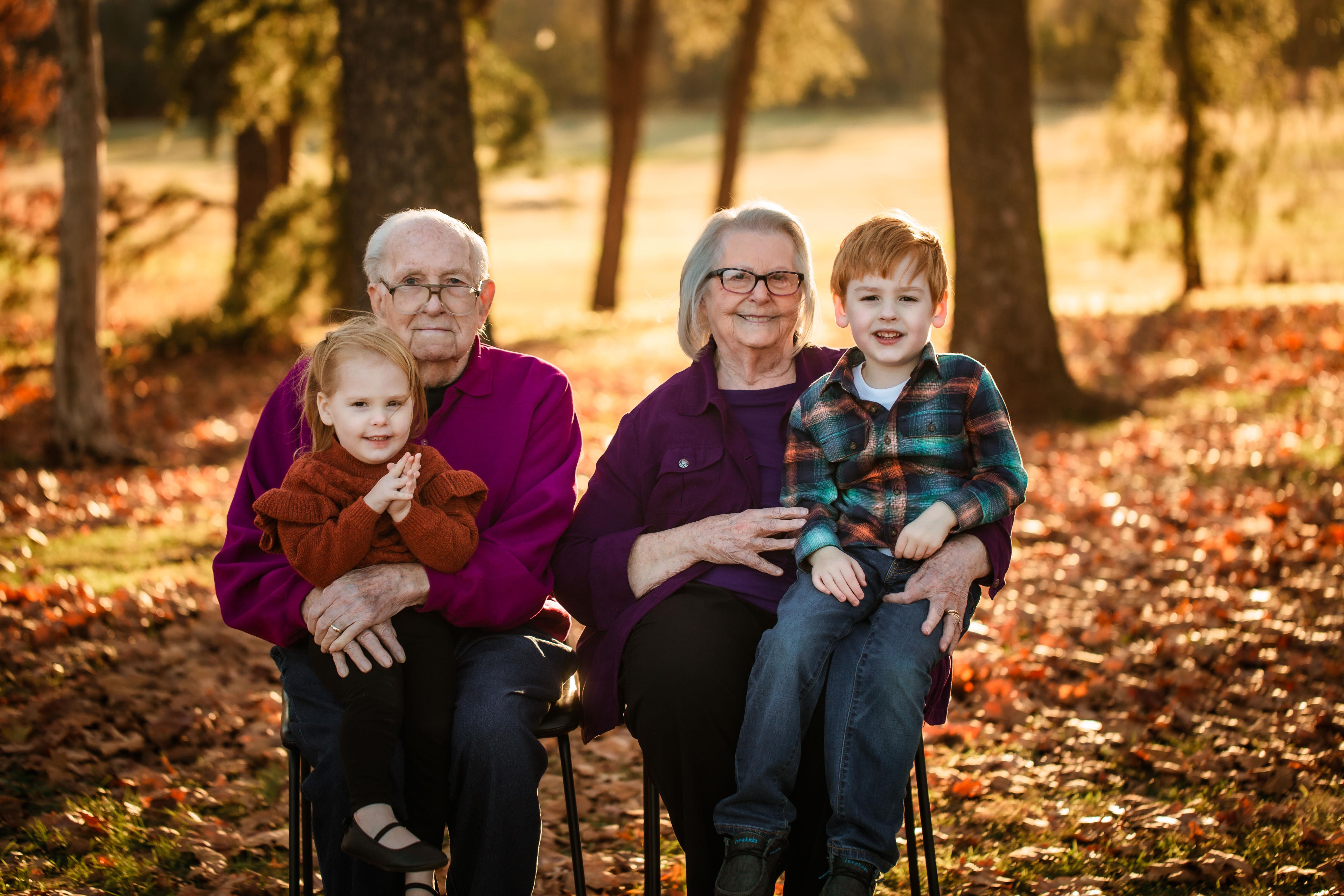 Grandparents sit with their grandchildren in a park filled with autumn leaves.