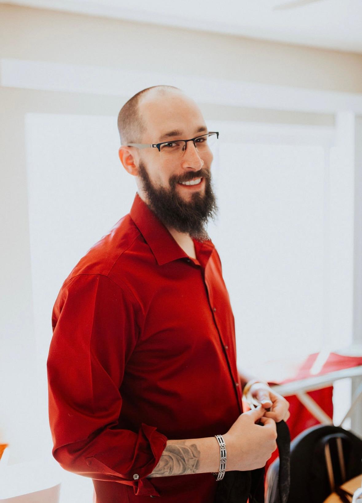 A man stands with a friendly expression, dressed in a red shirt, indoors with bright lighting.