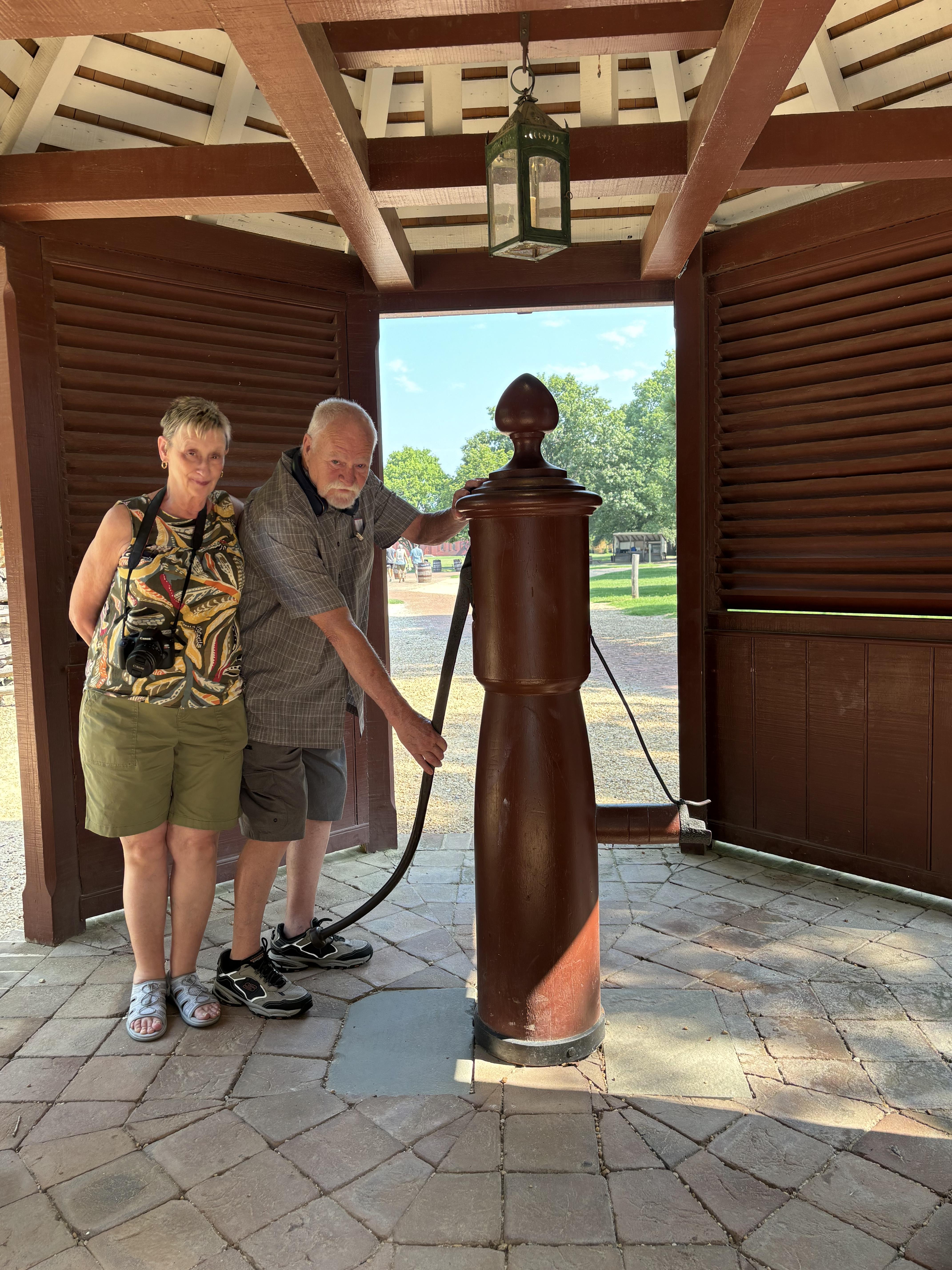 Two visitors explore a traditional water pump inside a wooden pavilion during daylight.