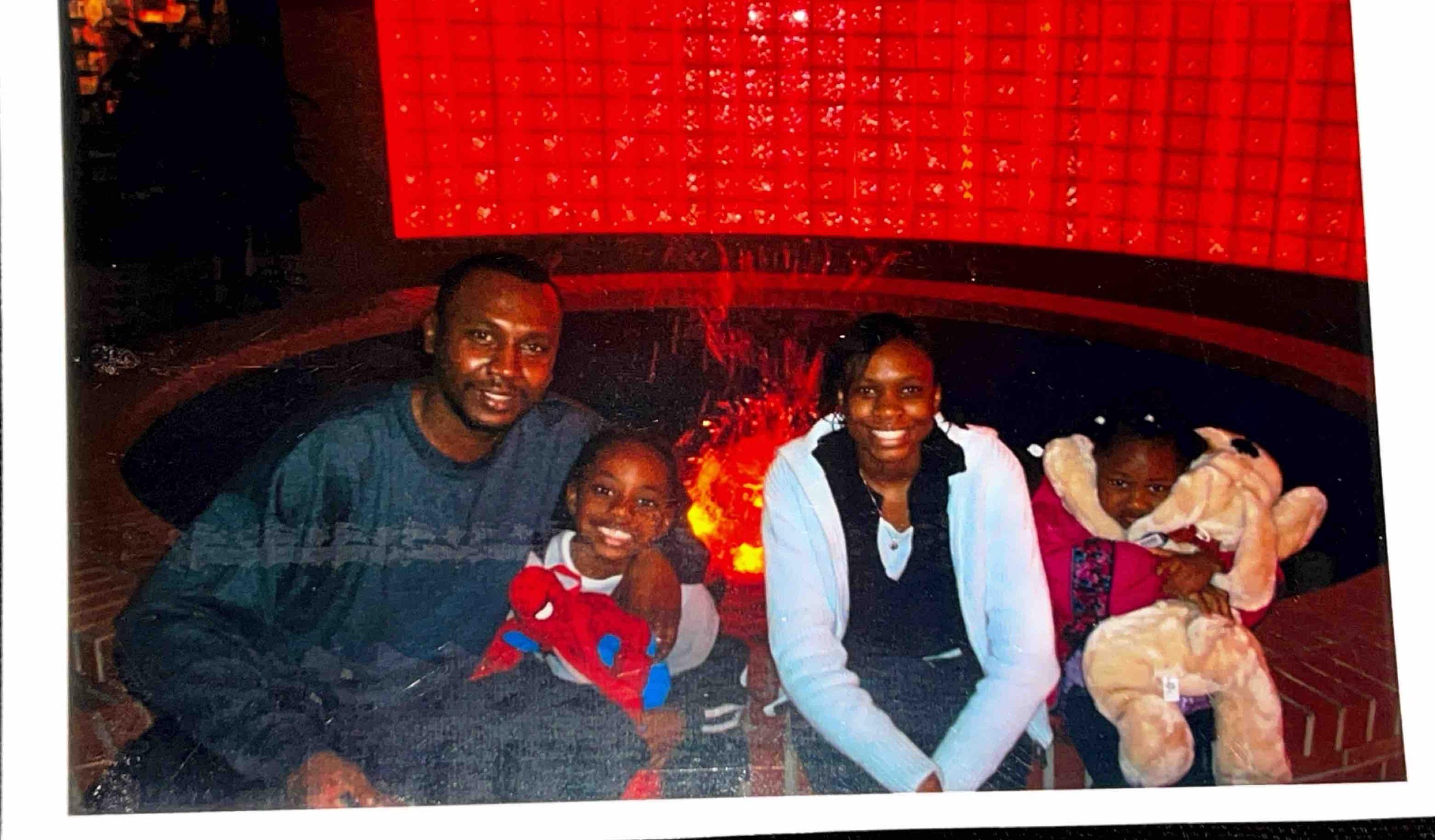 A family smiles cheerfully while sitting together near a colorful fountain in an indoor setting.