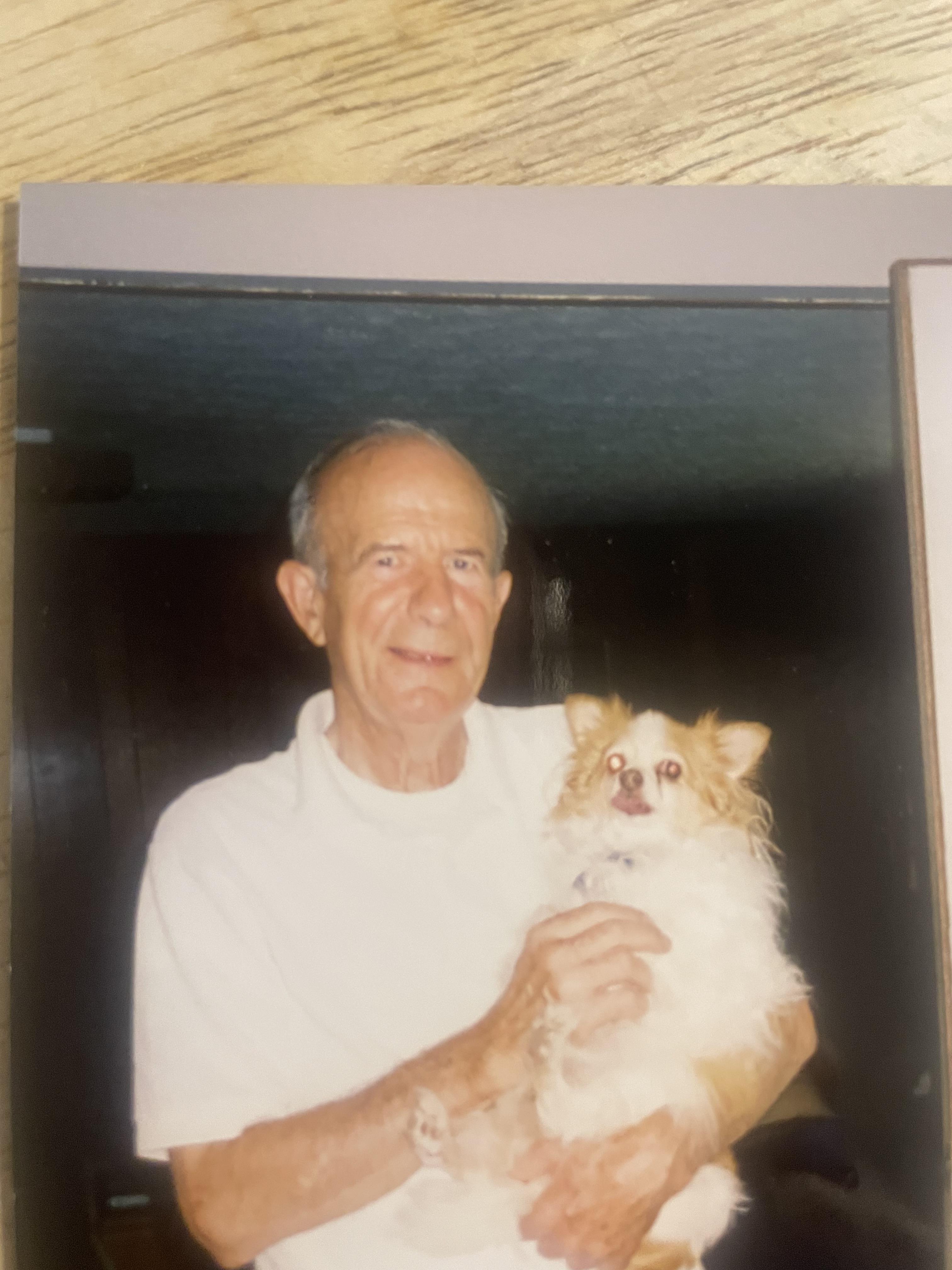 An elderly man smiles while holding a small fluffy dog in a warm living room.