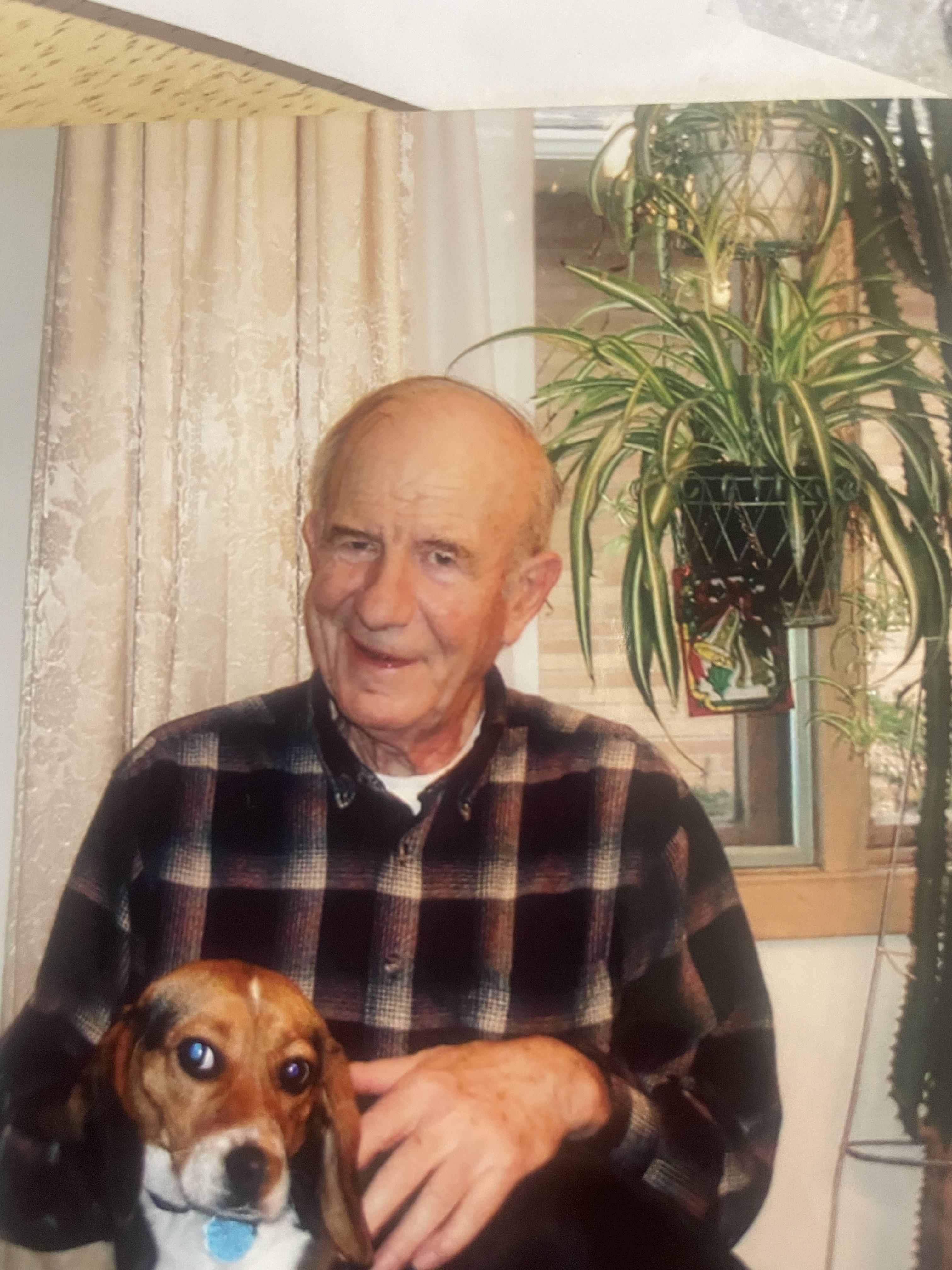 A smiling elderly man sits indoors with a small dog in his lap, surrounded by plants.