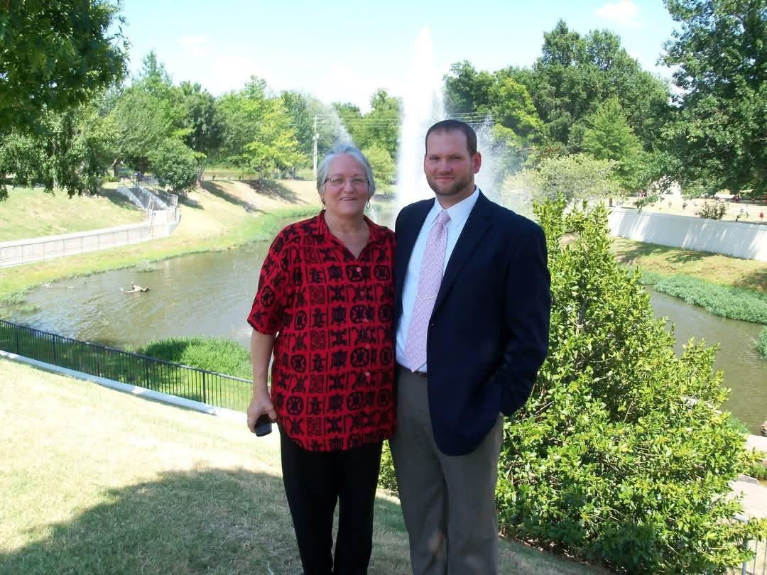 Two individuals smile together in a park, standing beside a water feature on a bright day.