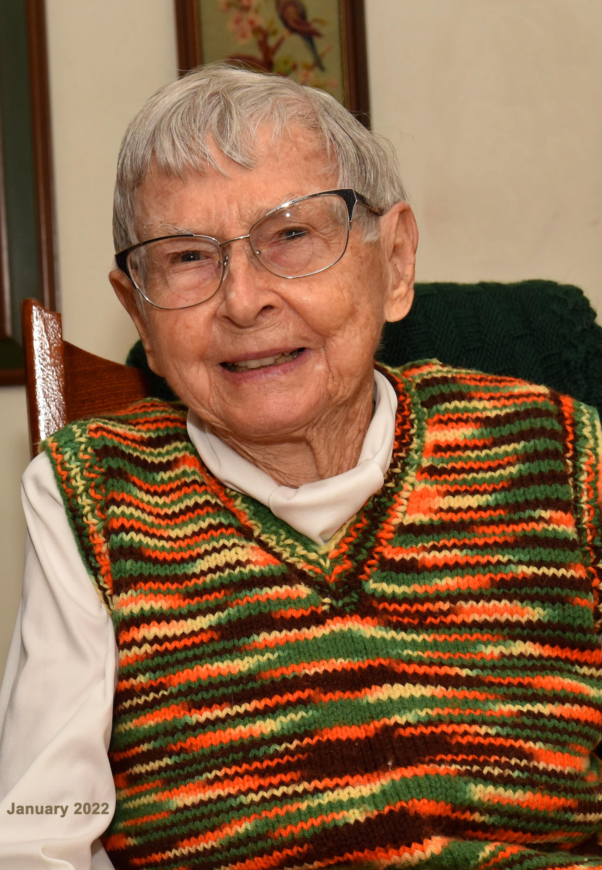 An elderly person sits comfortably, smiling warmly, dressed in a vibrant sweater in a bright room.