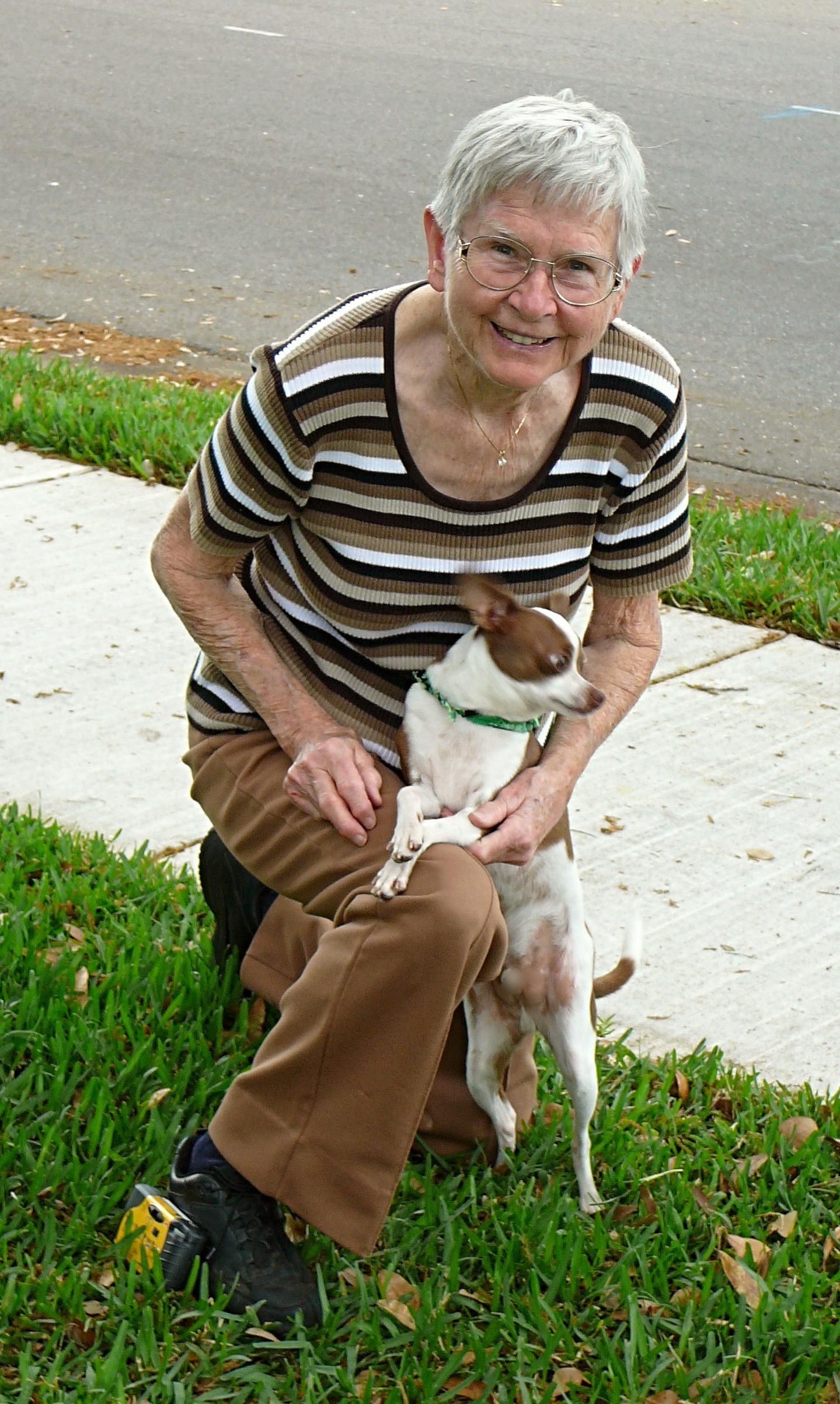 A joyful senior woman kneels on grass with her small dog, both enjoying a sunny afternoon together.
