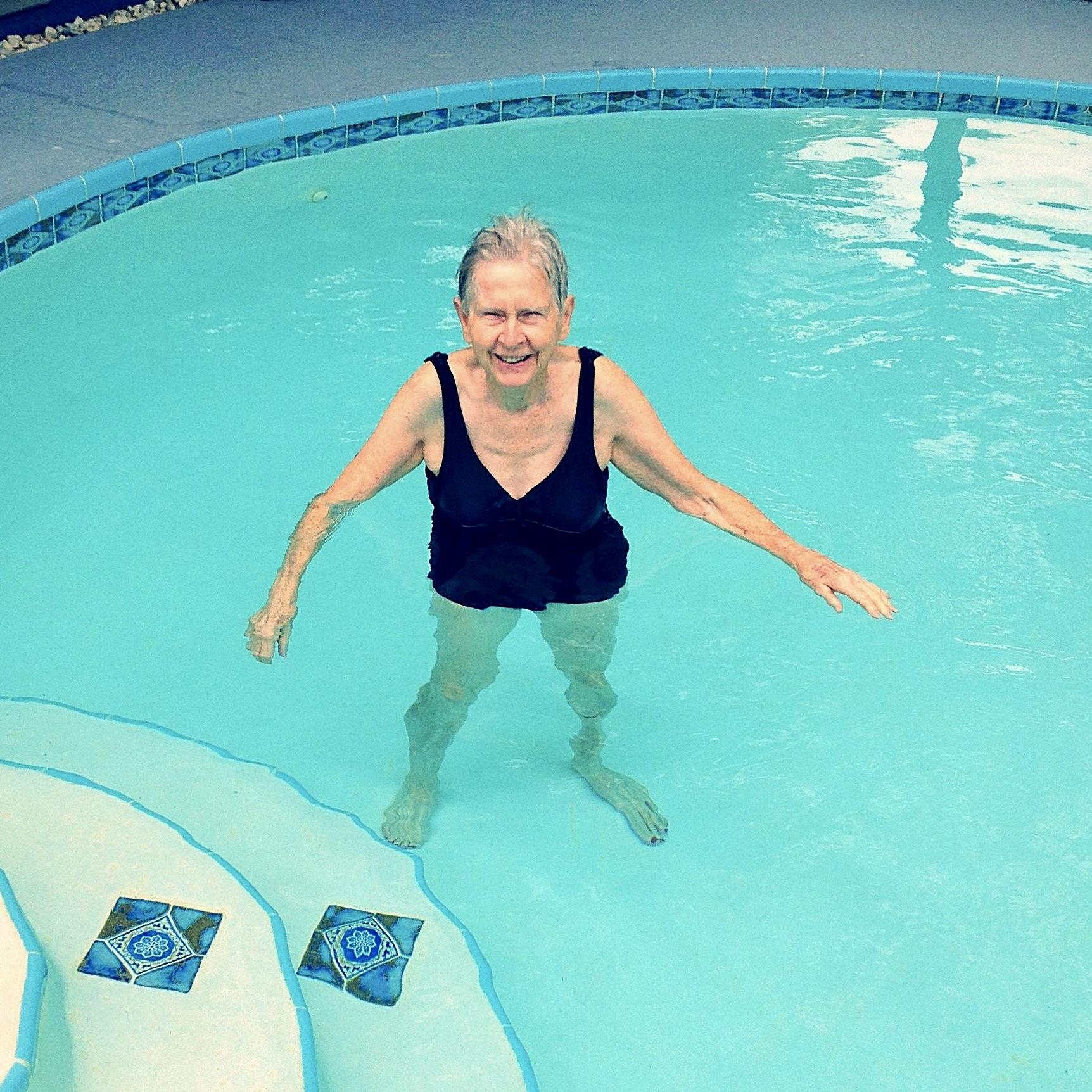 A cheerful elderly woman stands in a clear swimming pool, smiling and enjoying the warm water.