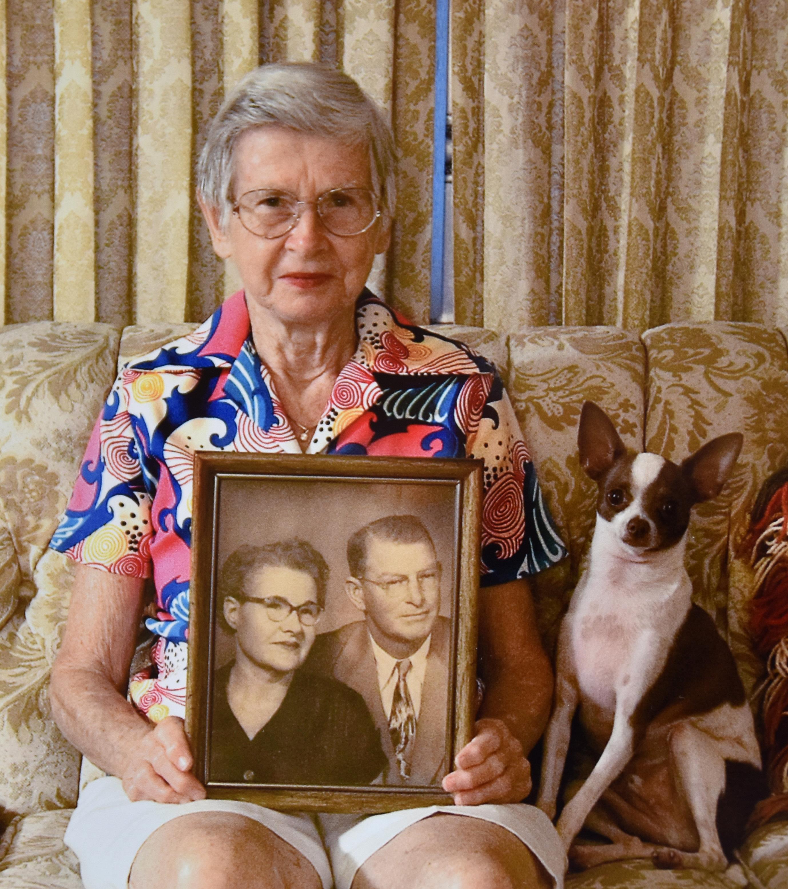 Senior woman proudly displays an old couple's portrait on a couch with a dog nearby.