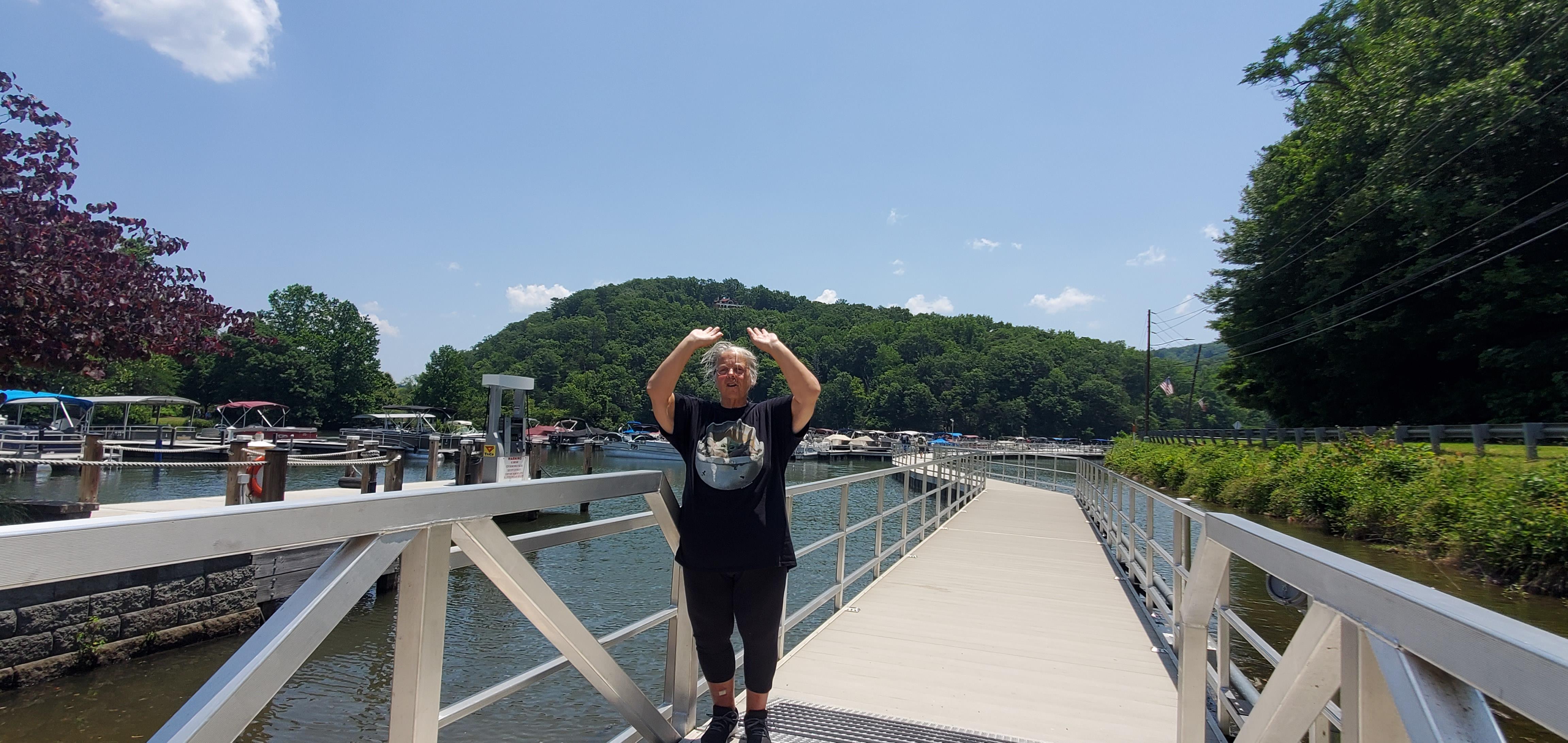Bright sunshine highlights a person joyfully celebrating on a lakeside dock surrounded by nature.