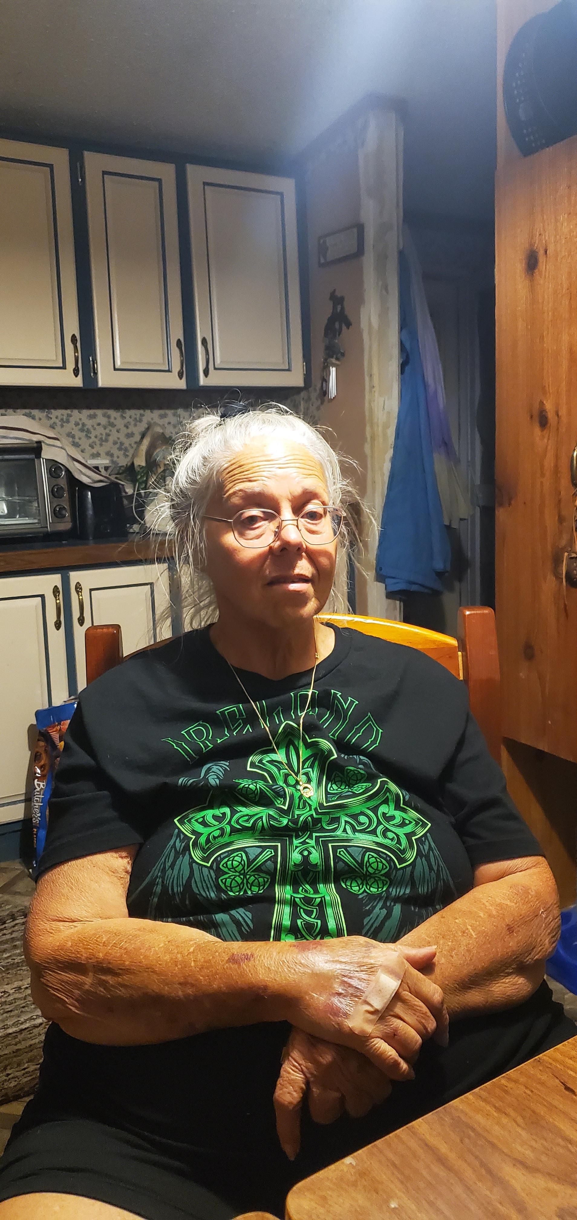 An elderly woman with gray hair sits quietly at a wooden table in a kitchen.