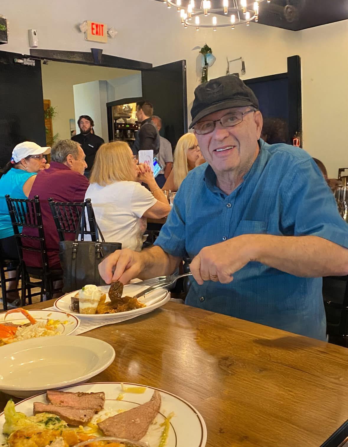 An elderly man is seated at a restaurant, smiling while enjoying a plate of food among friends.