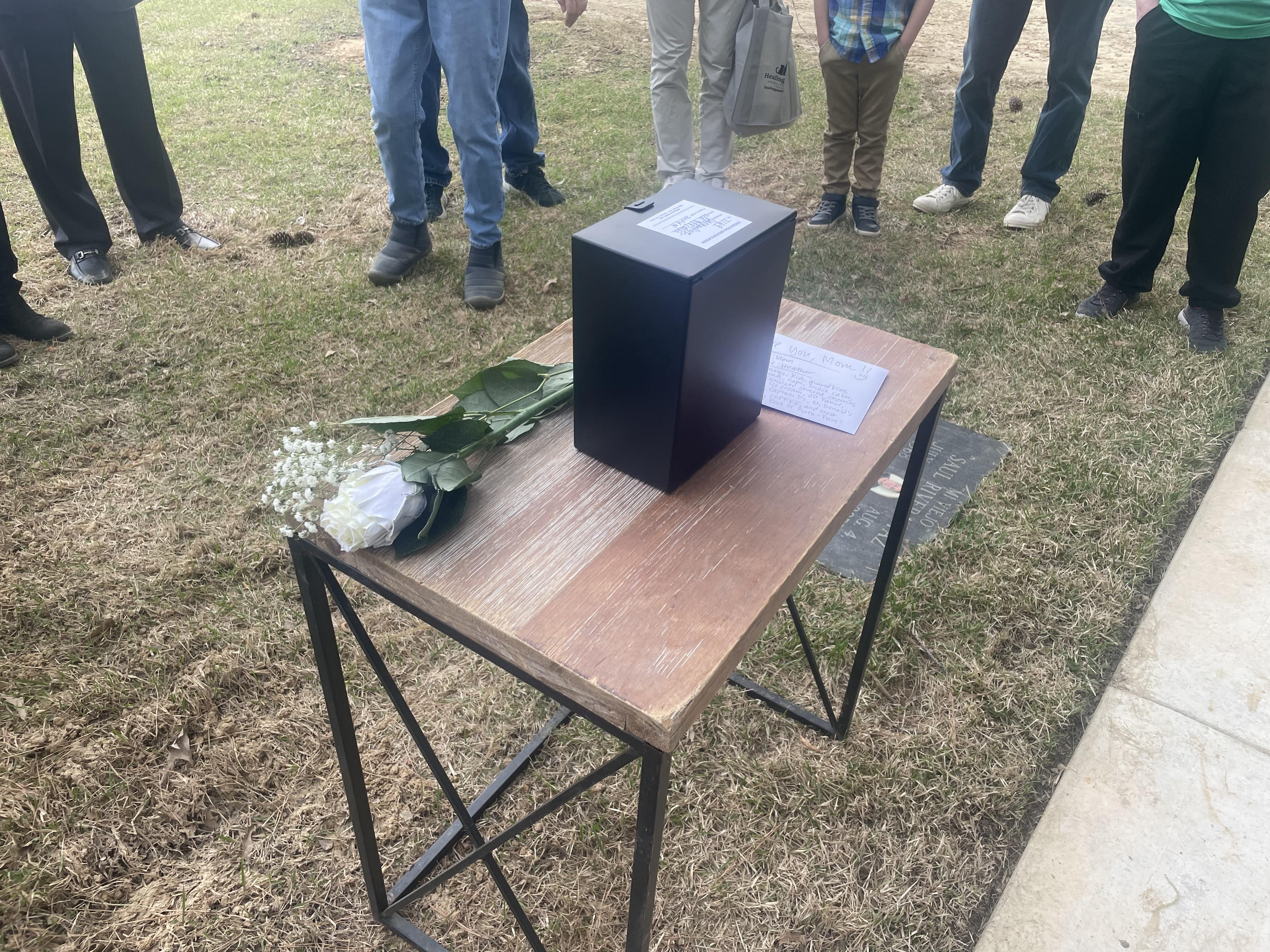 Relatives gather at a cemetery, honoring a loved one with flowers and a memory box.