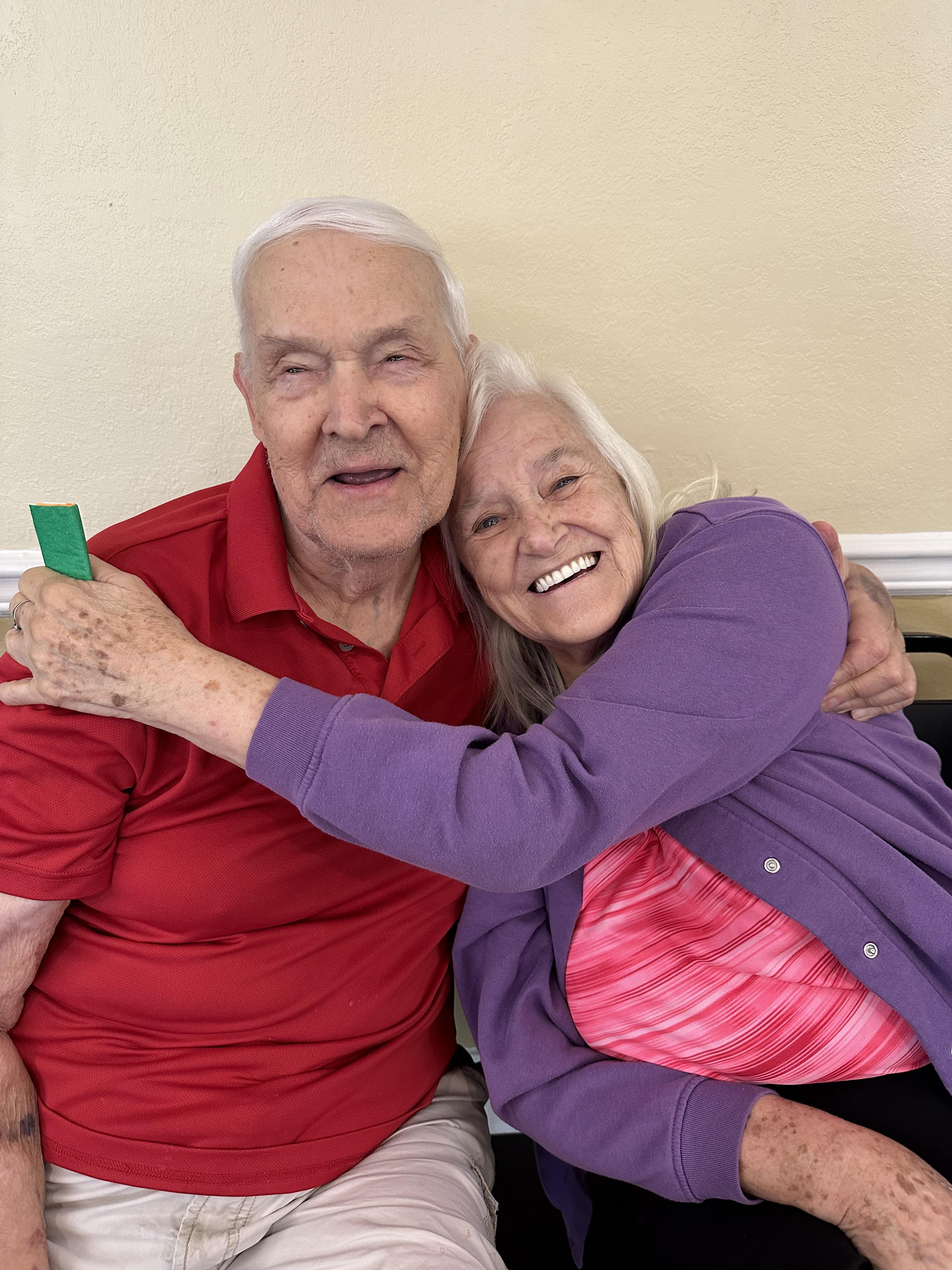 Two elderly individuals share a warm embrace and smile while seated in a comfortable space.