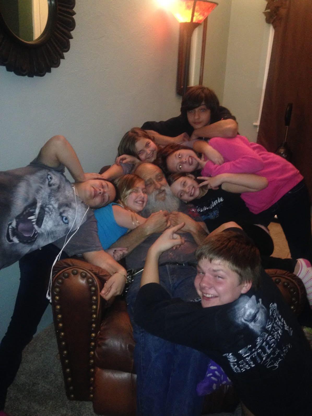 Children gather around an older man on a chair, all smiling and enjoying each other's company.