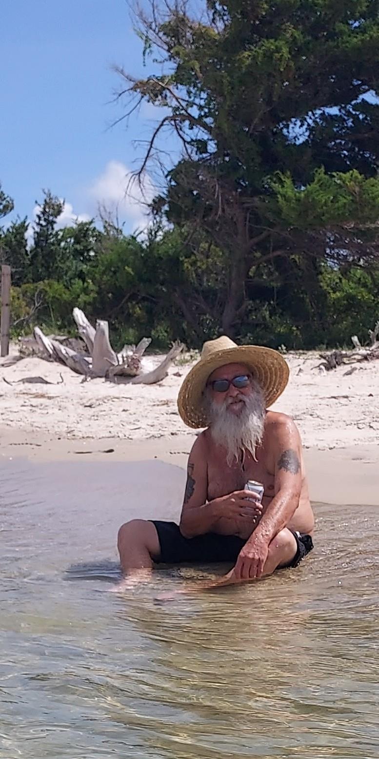 A man with a beard relaxes in shallow water on a sandy beach, soaking up the sun.