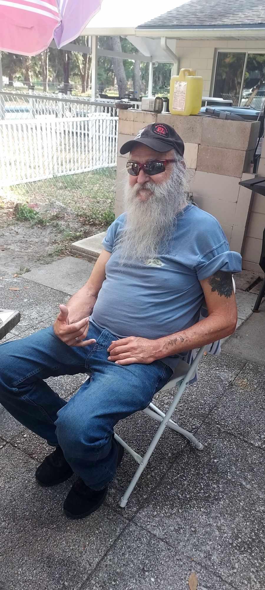 Elderly man with a long beard enjoys his time sitting outdoors, wearing casual clothes and a cap.