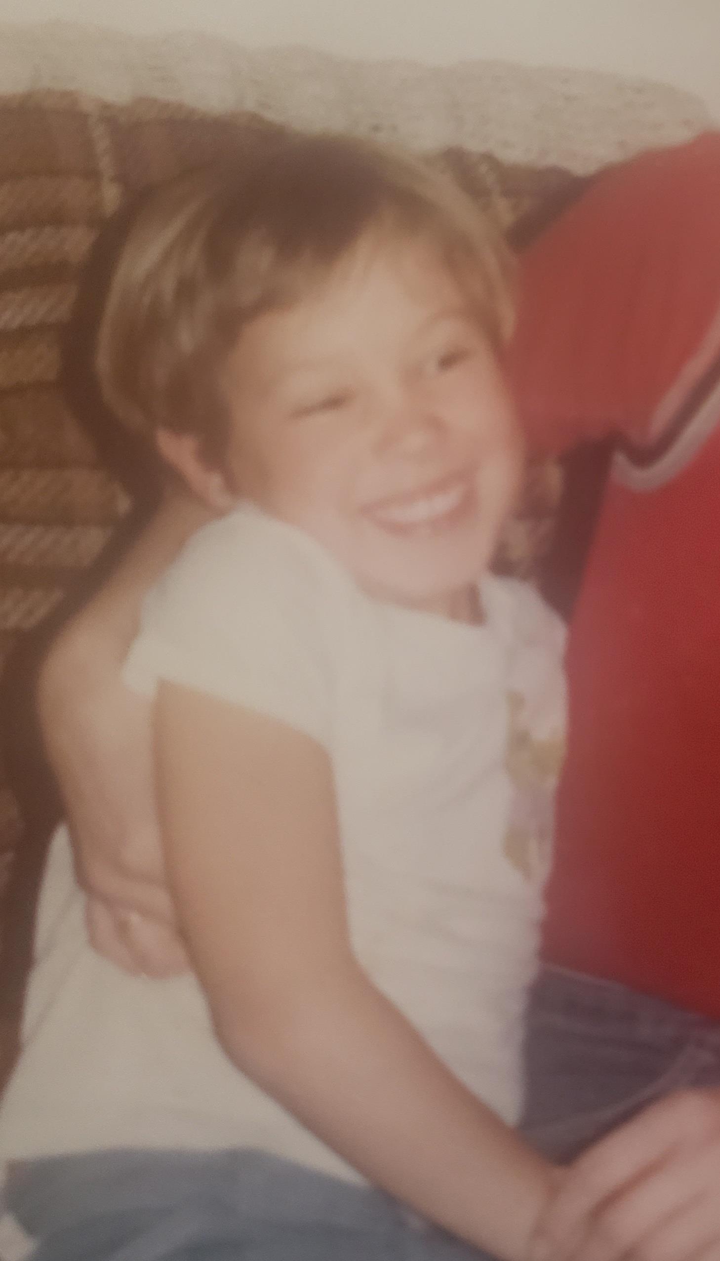 A young child with short hair beams joyfully while being embraced by an adult on a couch.