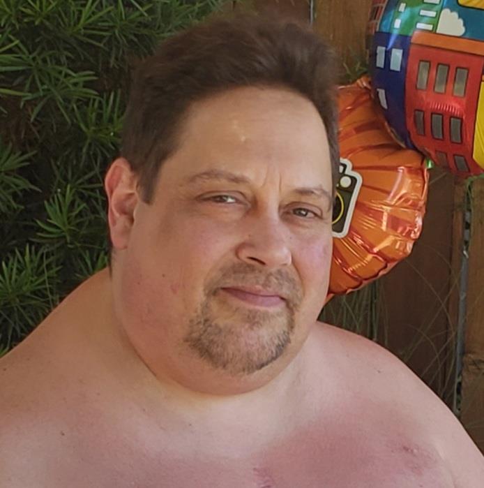 A man relaxes outside, smiling near colorful balloons in a garden on a sunny day.