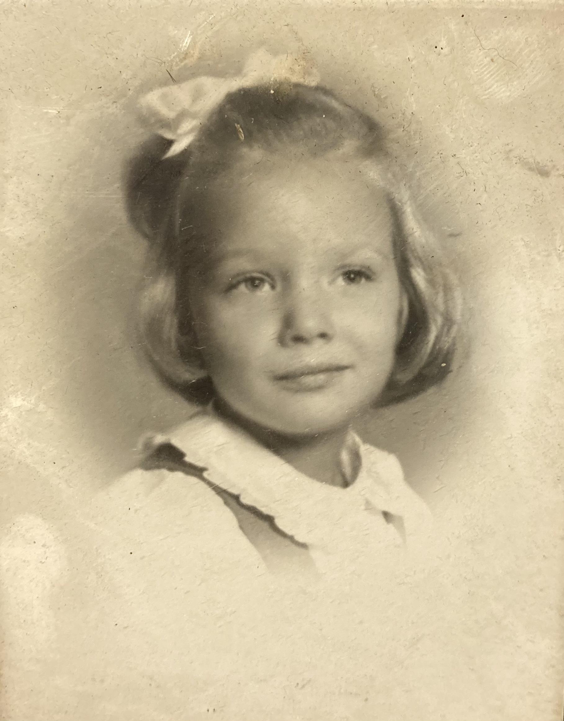 A girl with blonde hair and a bow looks directly at the camera, showcasing her innocence and charm.