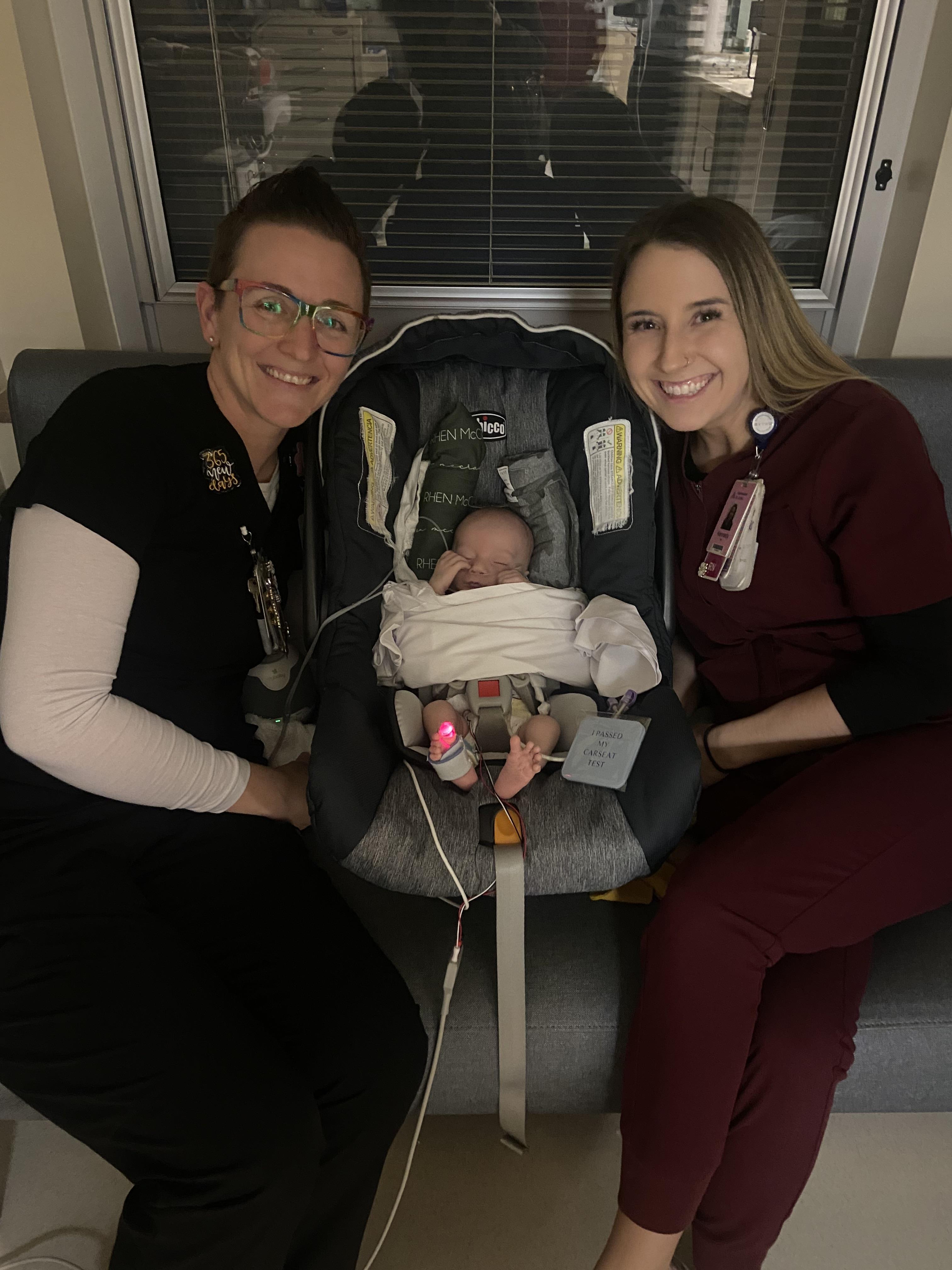 Two caregivers joyfully pose with a newborn in a warm, inviting room, capturing a special moment.