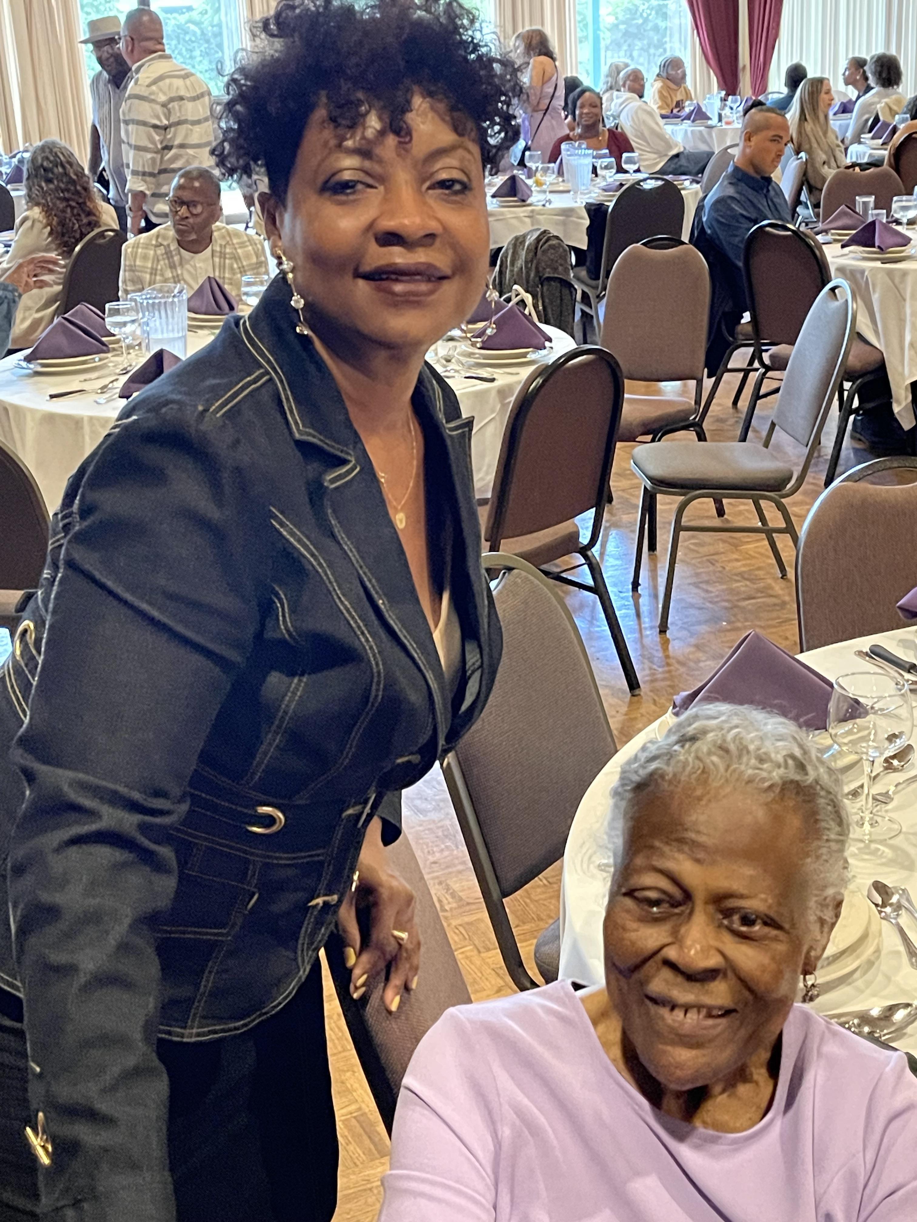 Two women smile warmly during a lively family event in a bustling hall filled with guests.