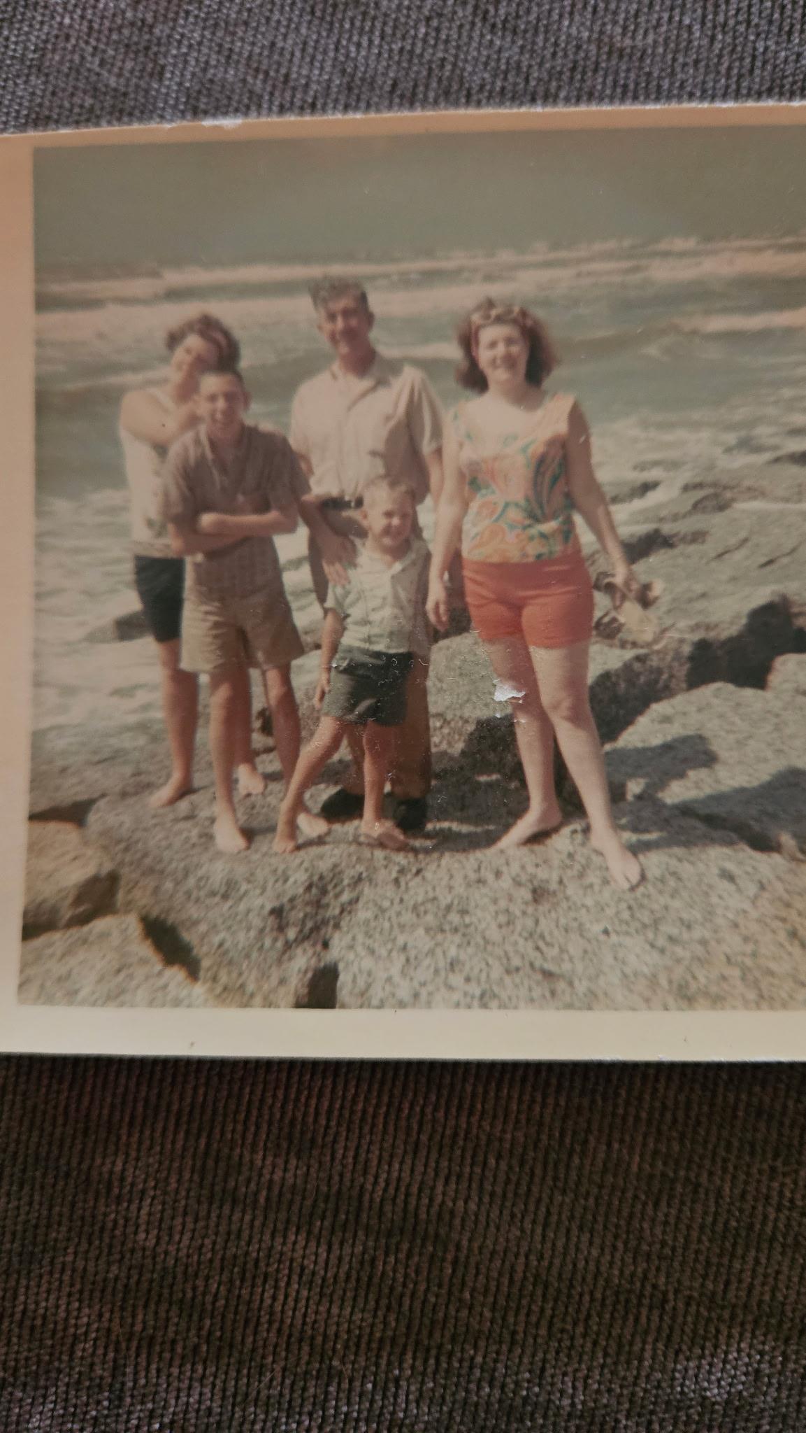 Family stands on the rocky beach with smiles, dressed in summer clothes enjoying the day together.