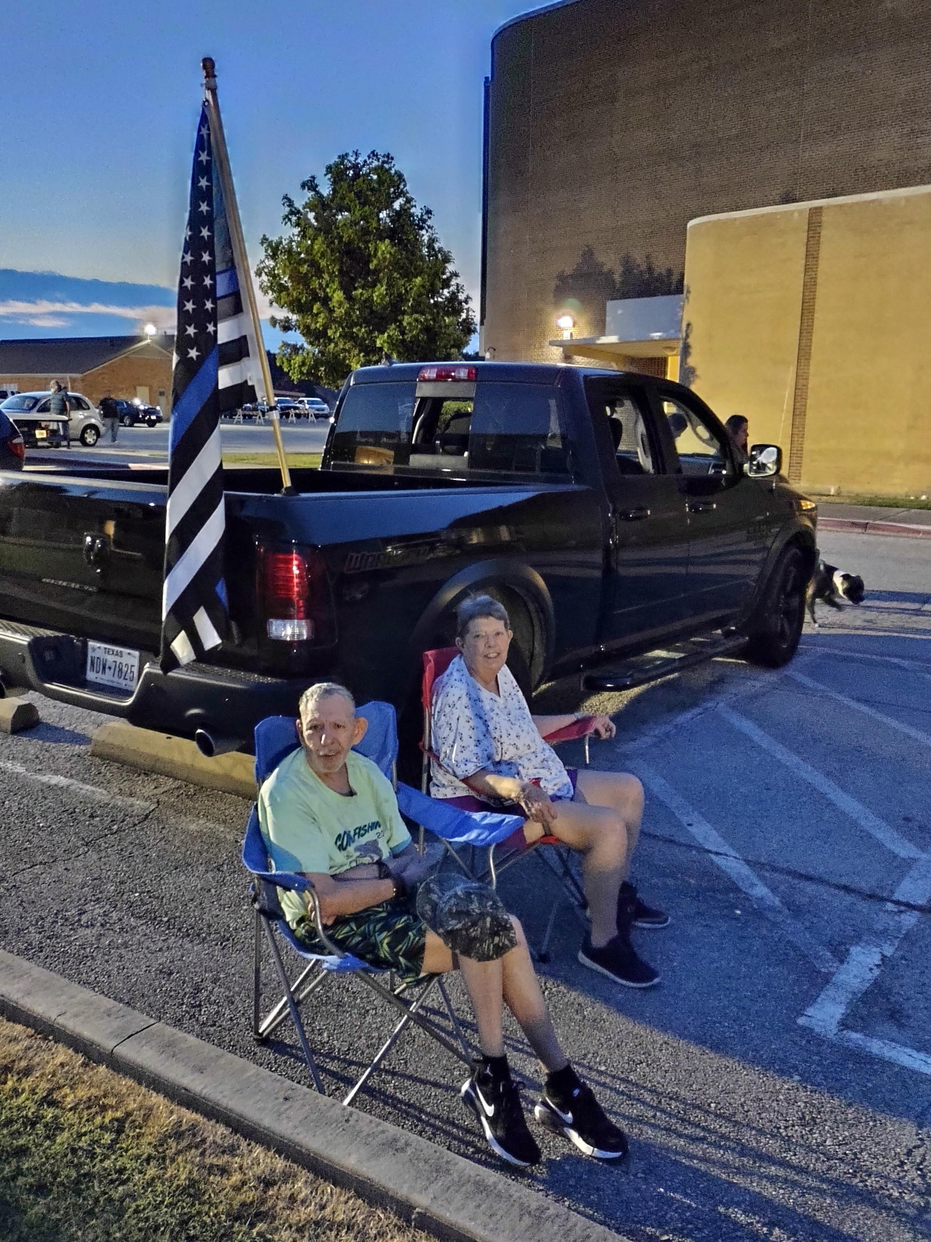 Two older men sit in chairs near a pickup truck with an American flag while enjoying the evening.