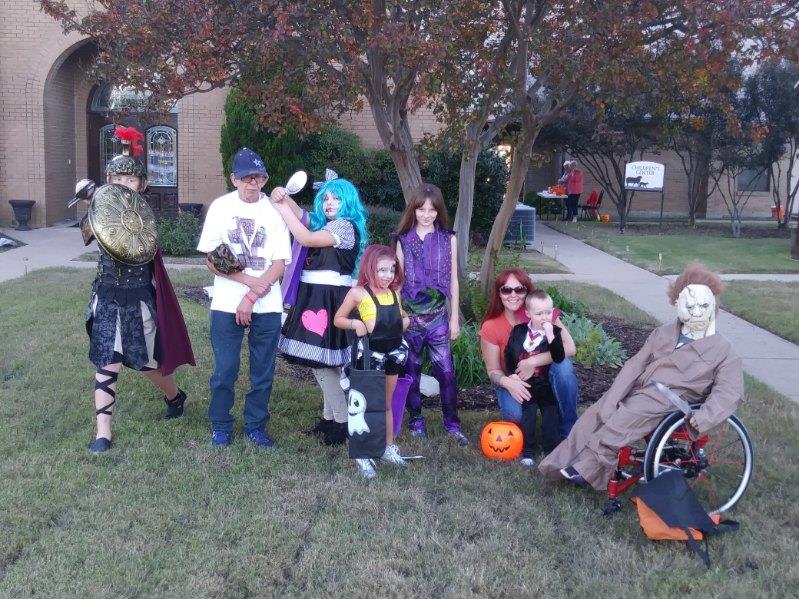 Various people in costumes gather outside near a tree to celebrate Halloween festivities.