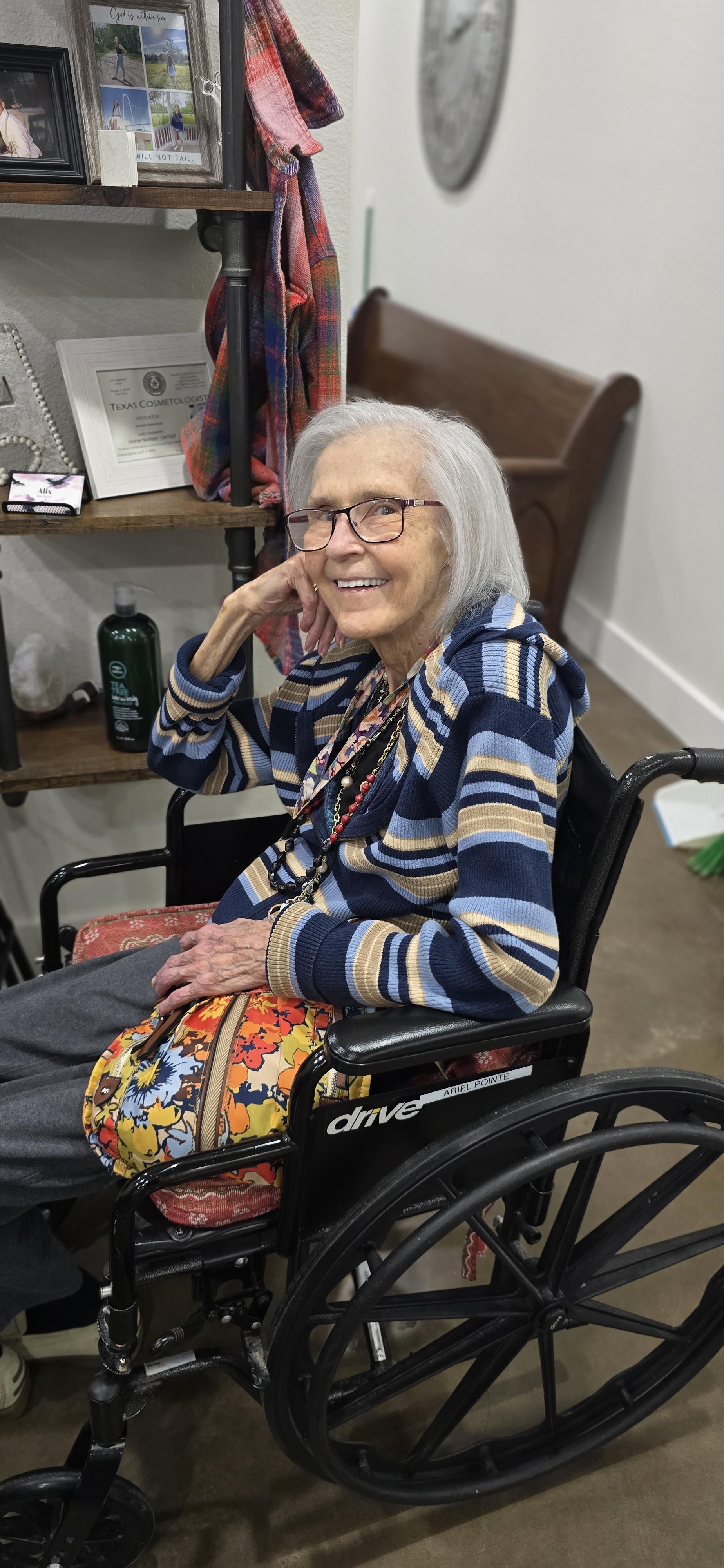 An elderly woman sits in a wheelchair, smiling and wearing glasses, in a warm, inviting atmosphere.