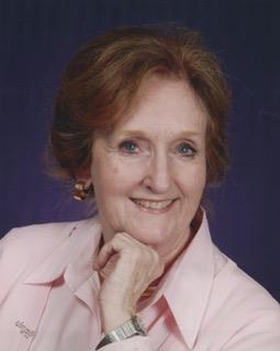 A woman smiling gently with brown hair styled, dressed in a light pink blouse, exuding warmth.