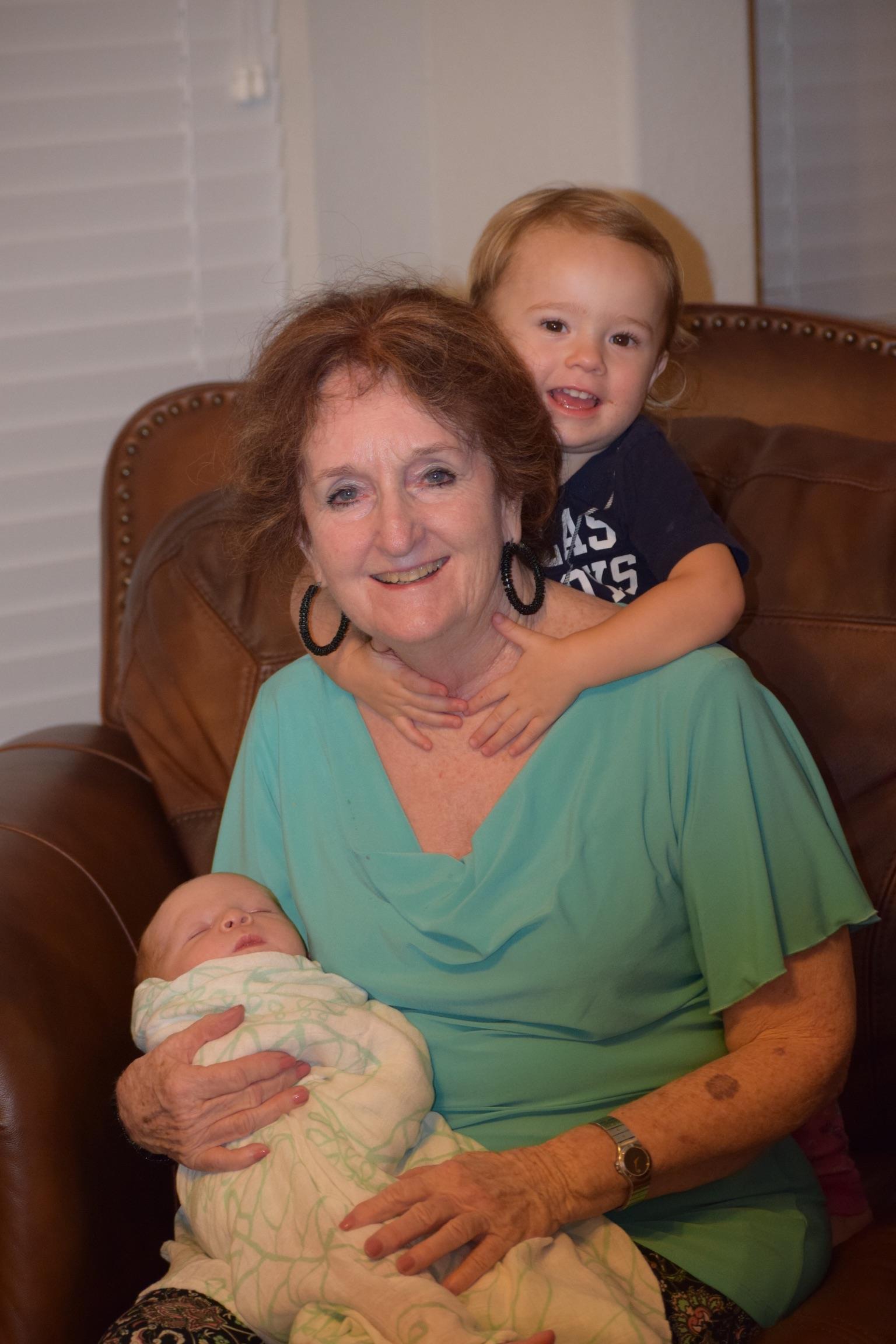 A grandmother holds a baby while her older grandchild hugs her from behind, smiling happily.