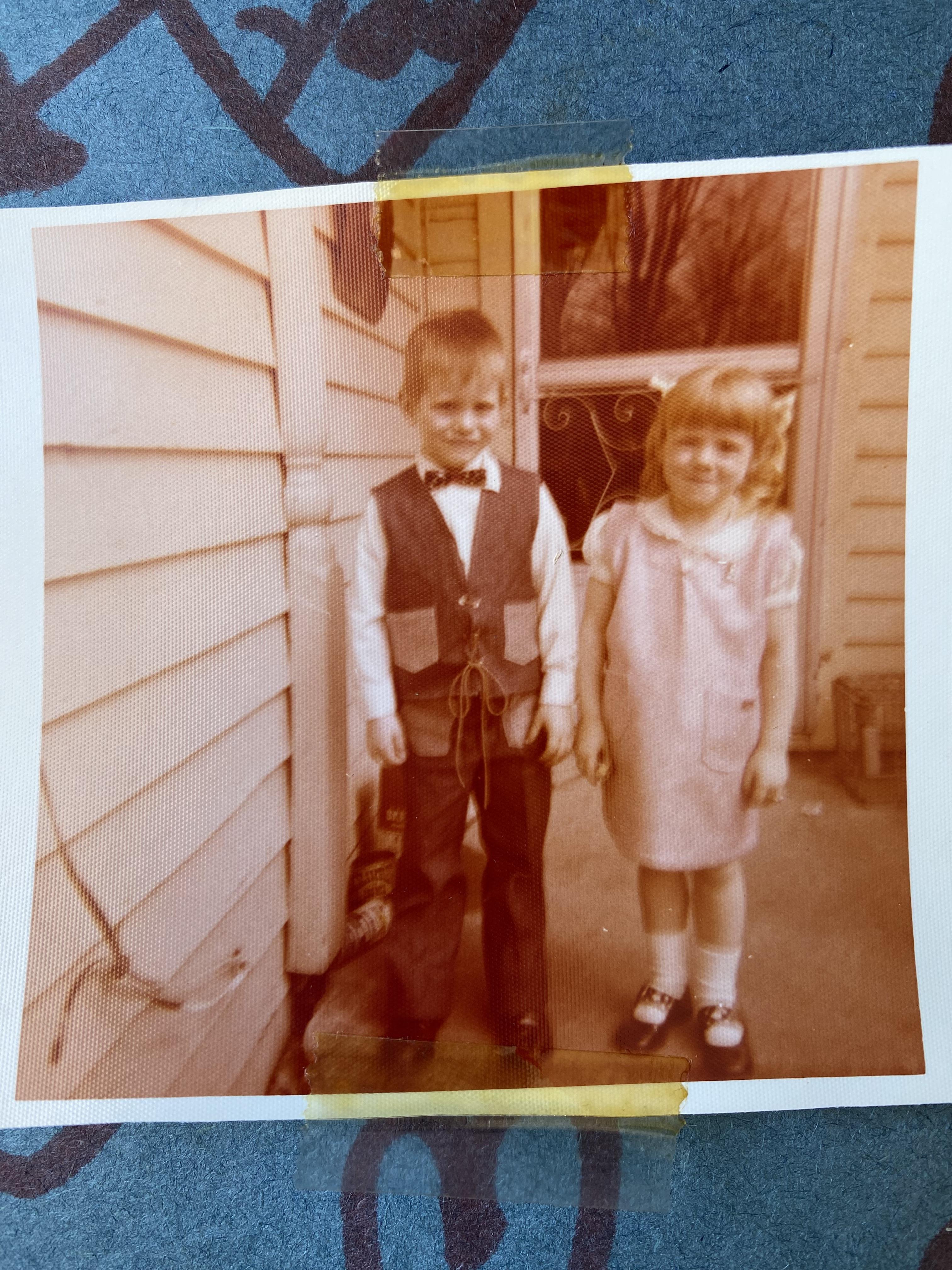 Two children stand together on a porch, dressed in vintage attire and smiling happily.
