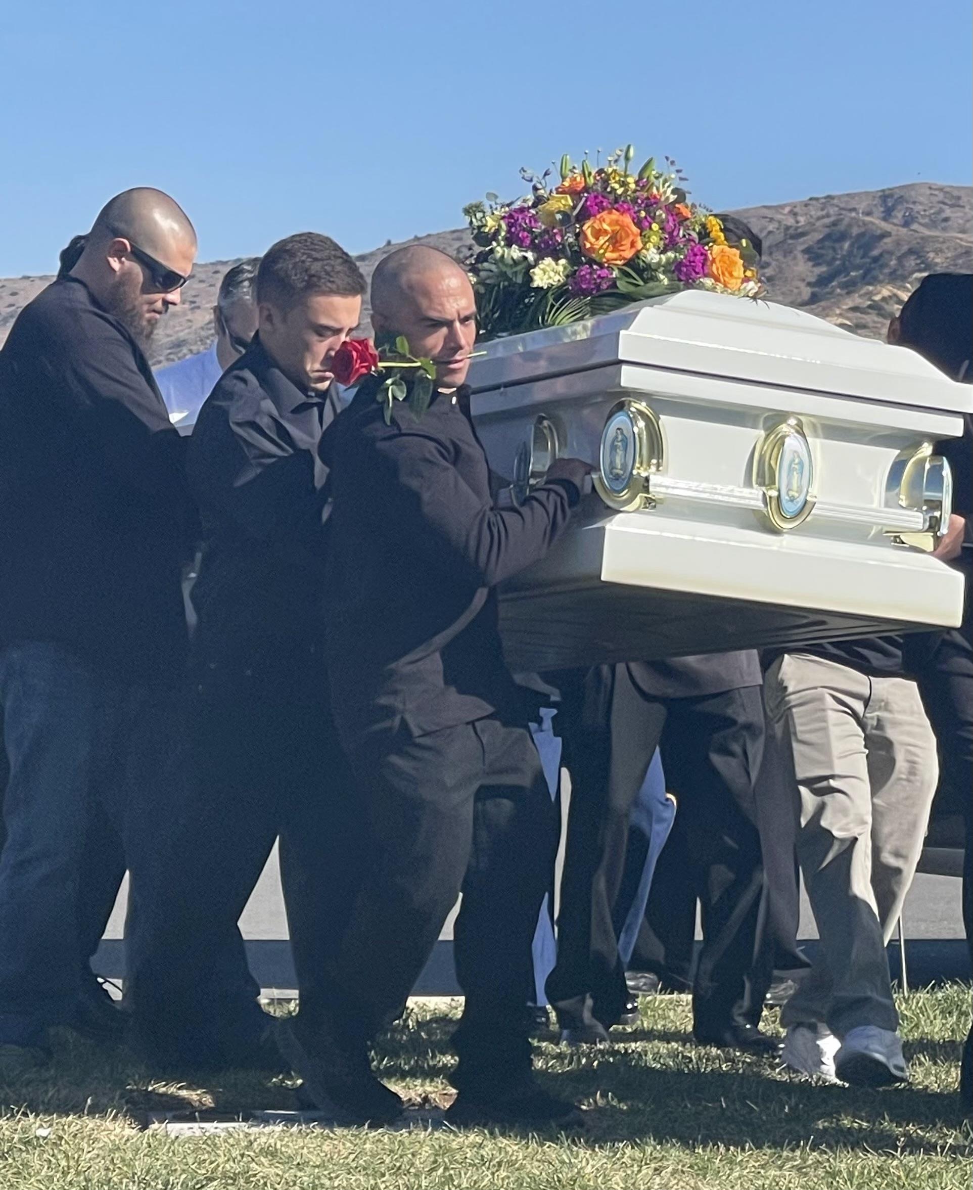 Mourners support each other while carrying a casket adorned with flowers at a burial.