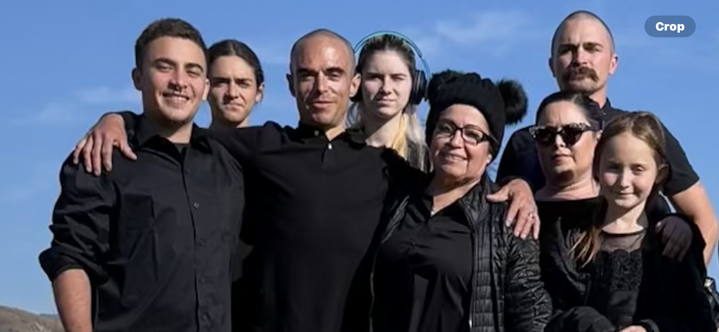 A diverse group of family members poses together outdoors, wearing black outfits, under a blue sky.