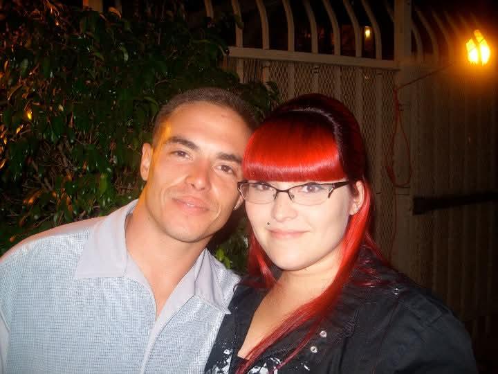 A couple poses closely with smiles, surrounded by evening lights and greenery.
