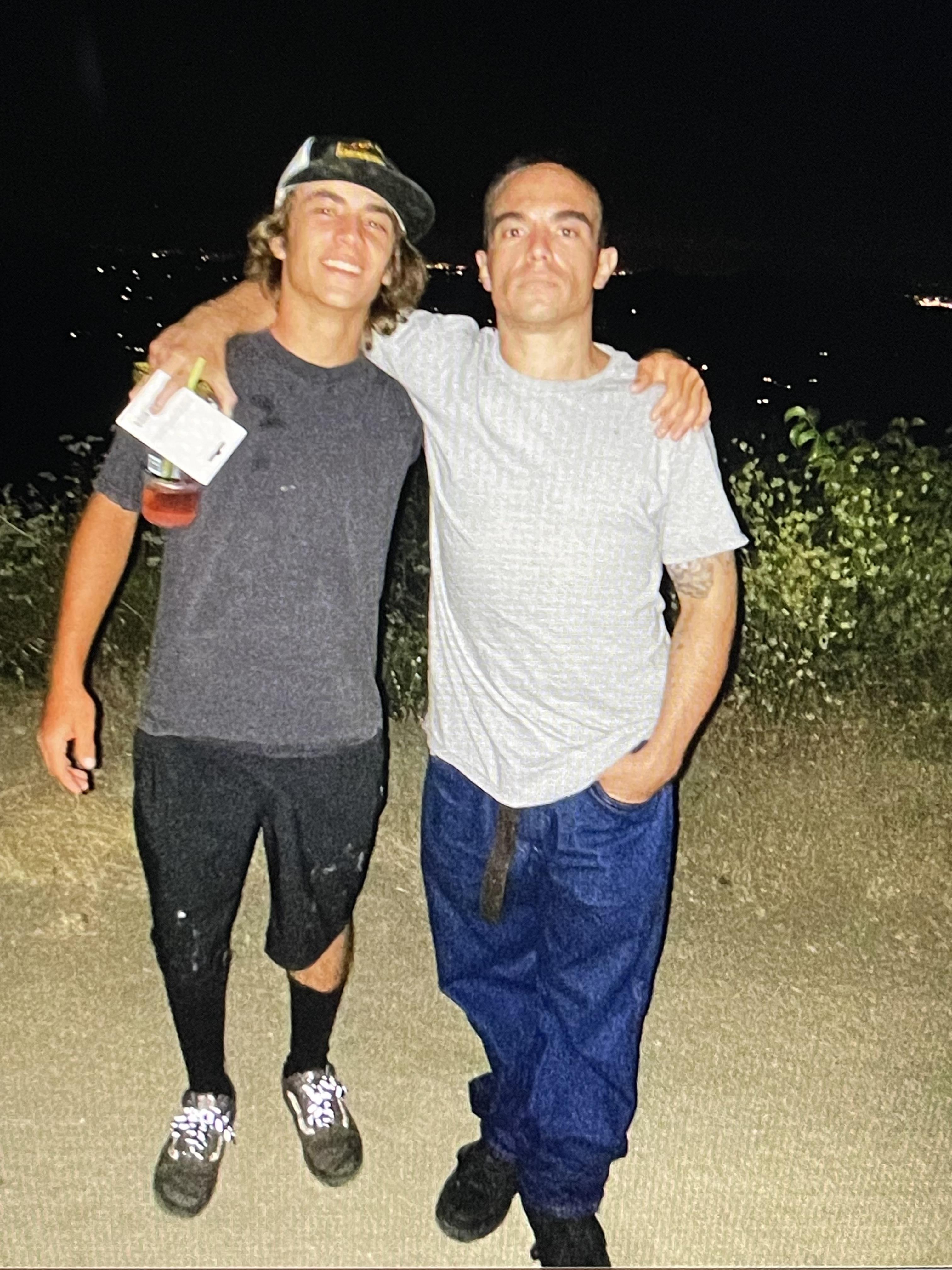 Two friends stand together at night, smiling and holding drinks by a calm waterway.