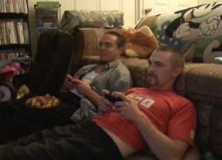 Two friends sit on a couch, engaged in a gaming session with snacks around.