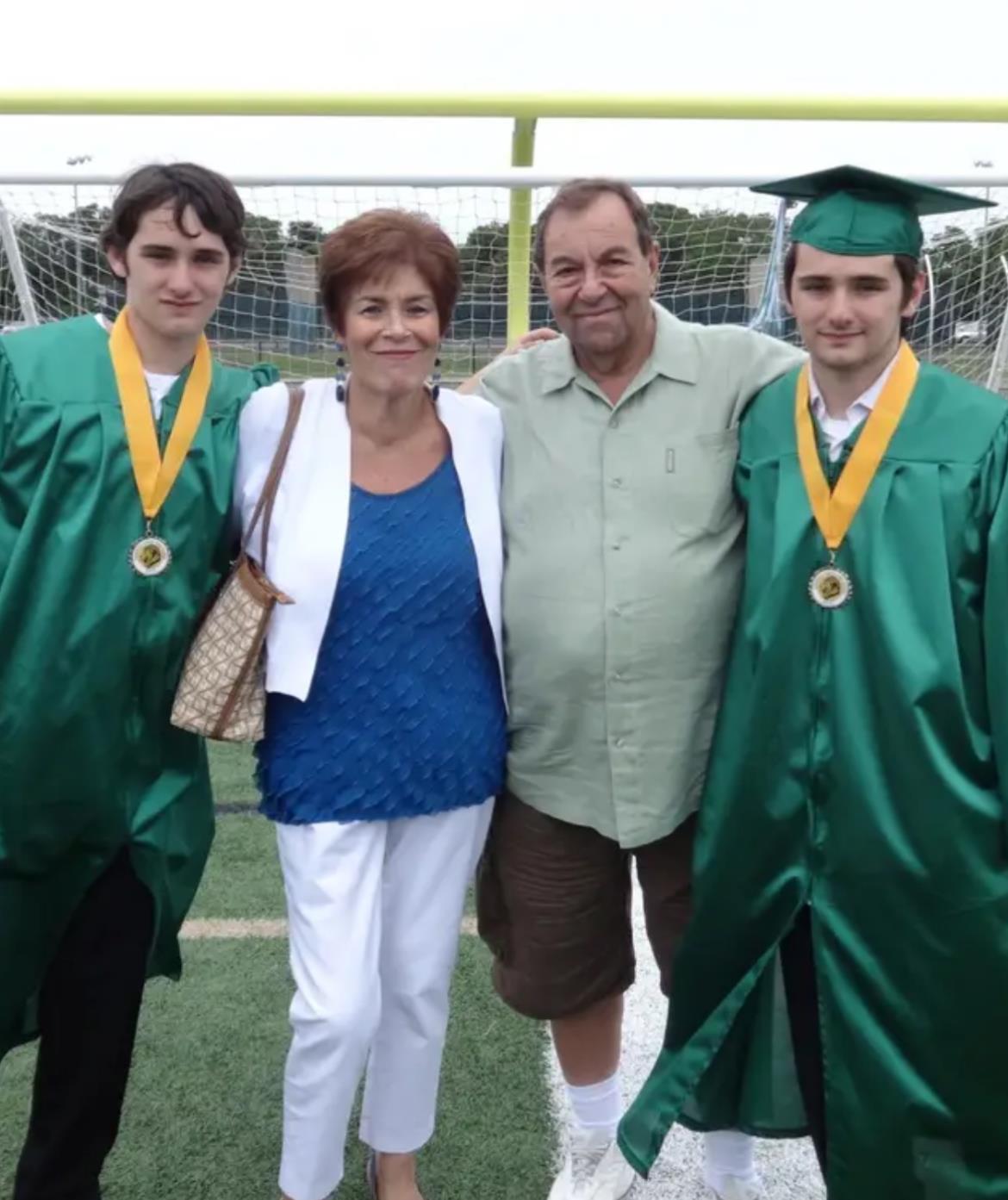 Family gathers on a sports field to celebrate the graduation of two students in caps and gowns.