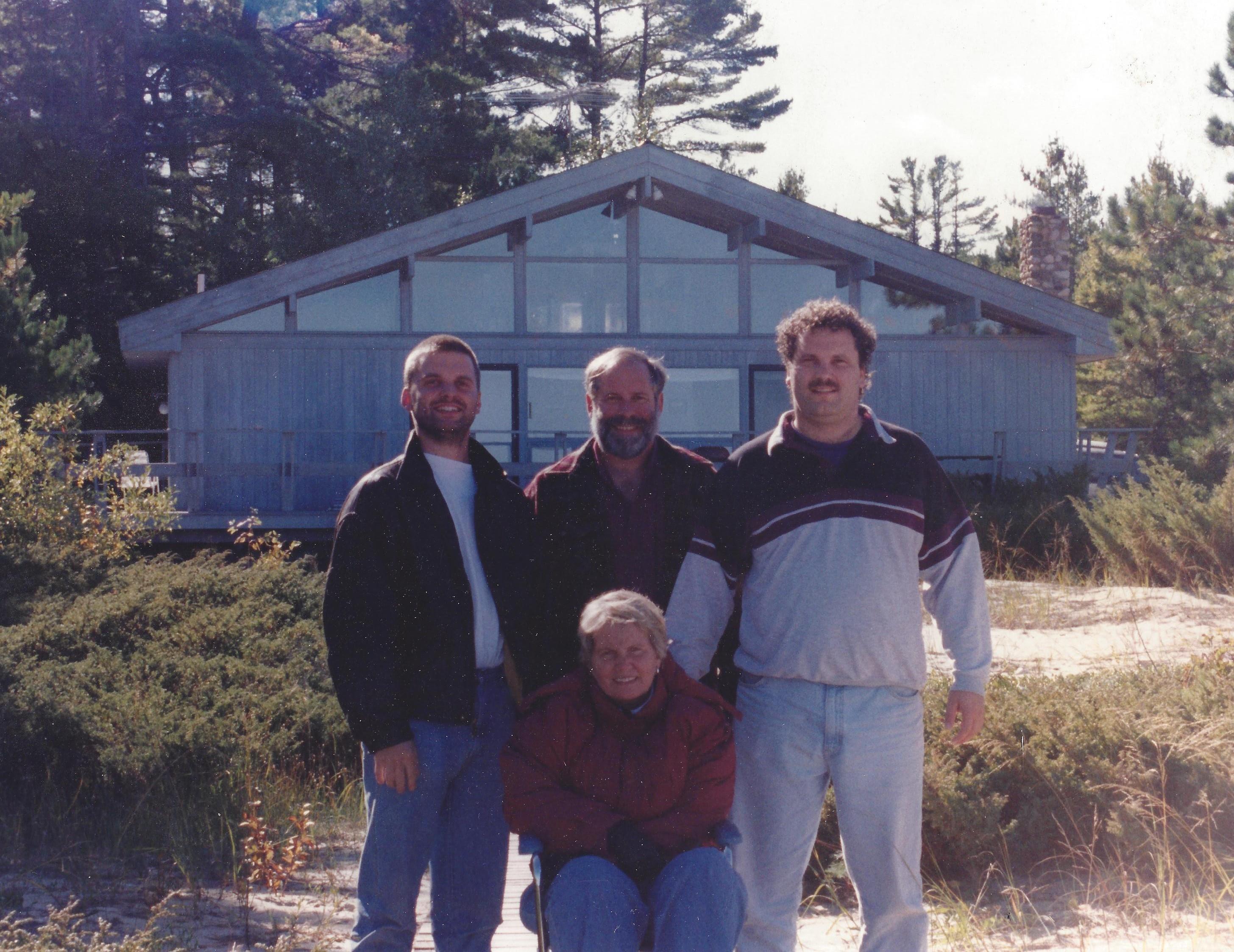 Three men stand with a child in front of a rustic cabin set in a forest during daylight.