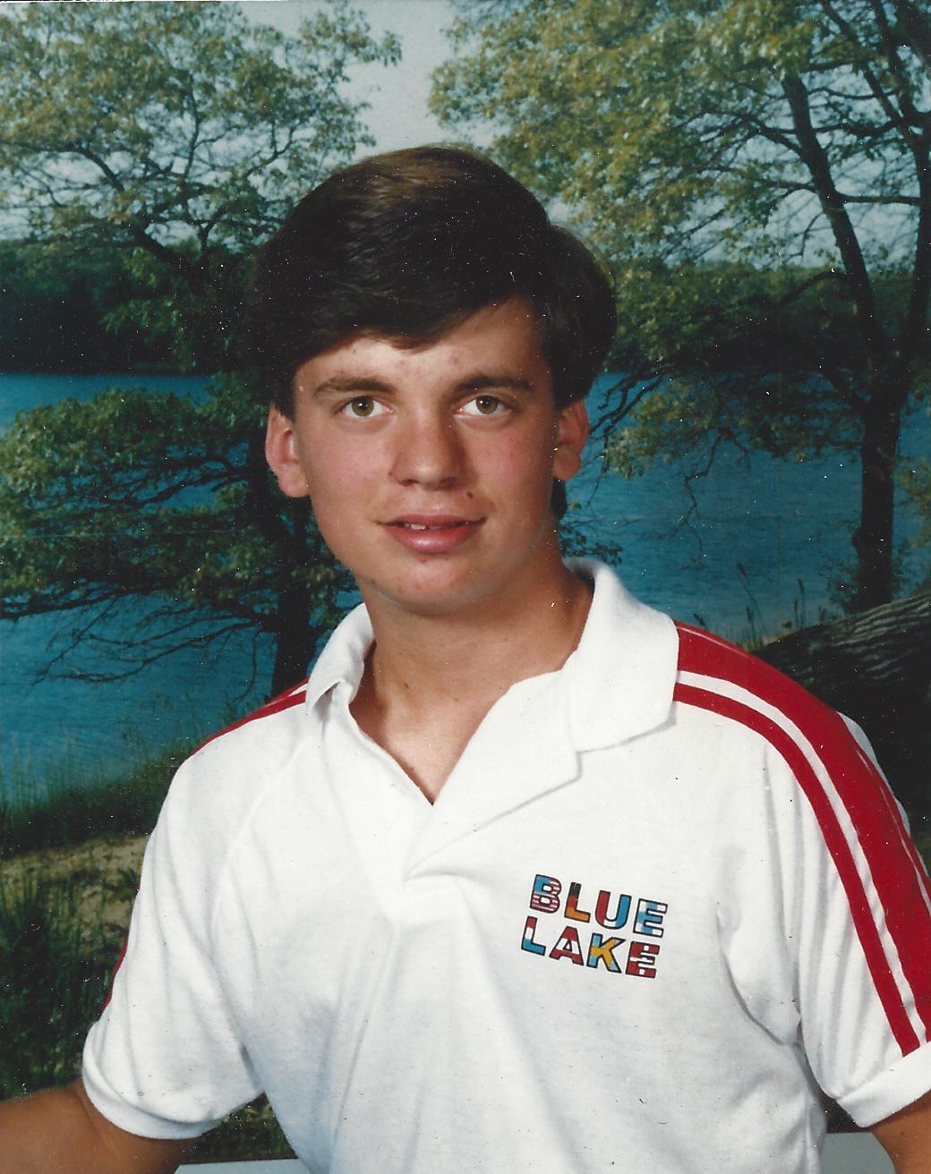 A young man smiles for the camera near a serene lake surrounded by trees on a sunny day.