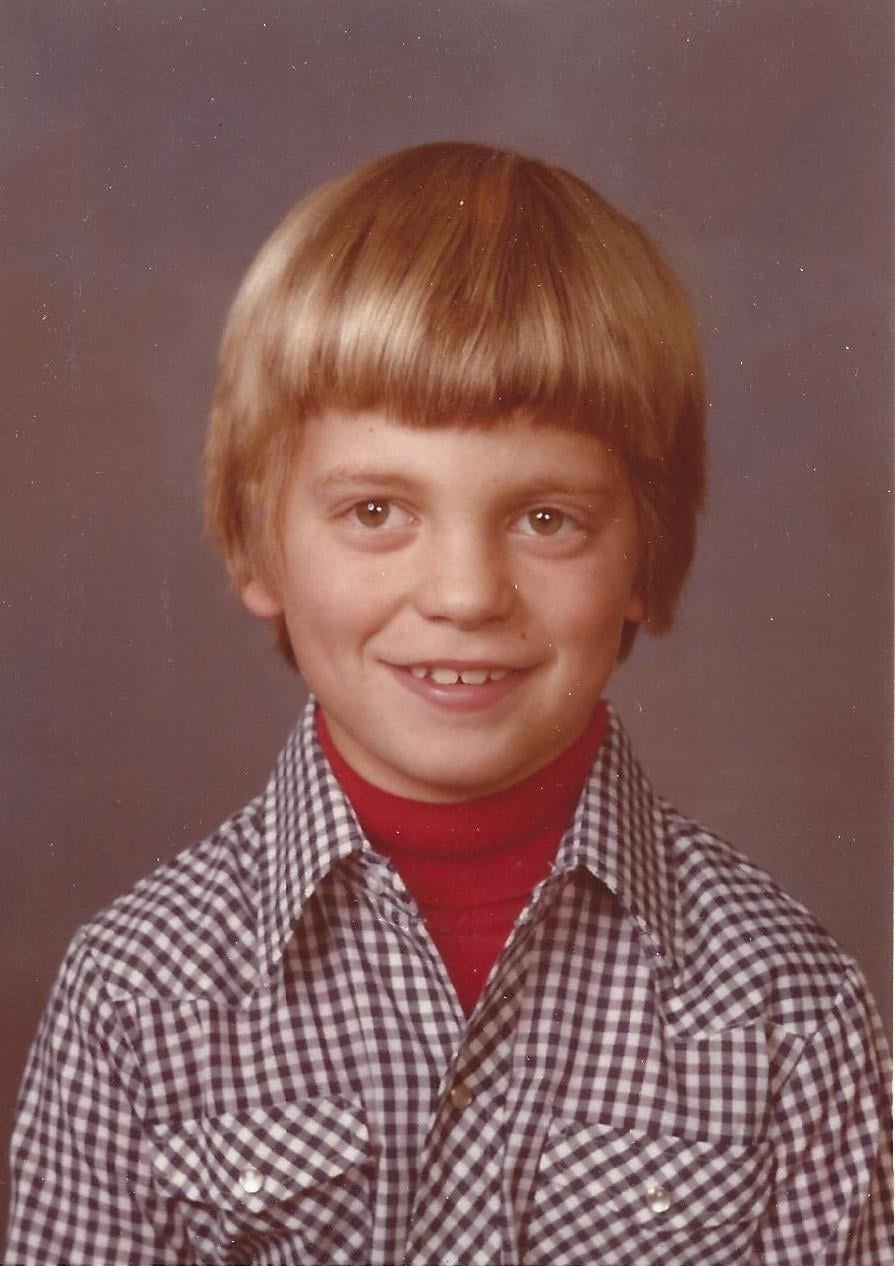 A cheerful young boy with blonde hair is smiling while wearing a checkered shirt and red turtleneck.