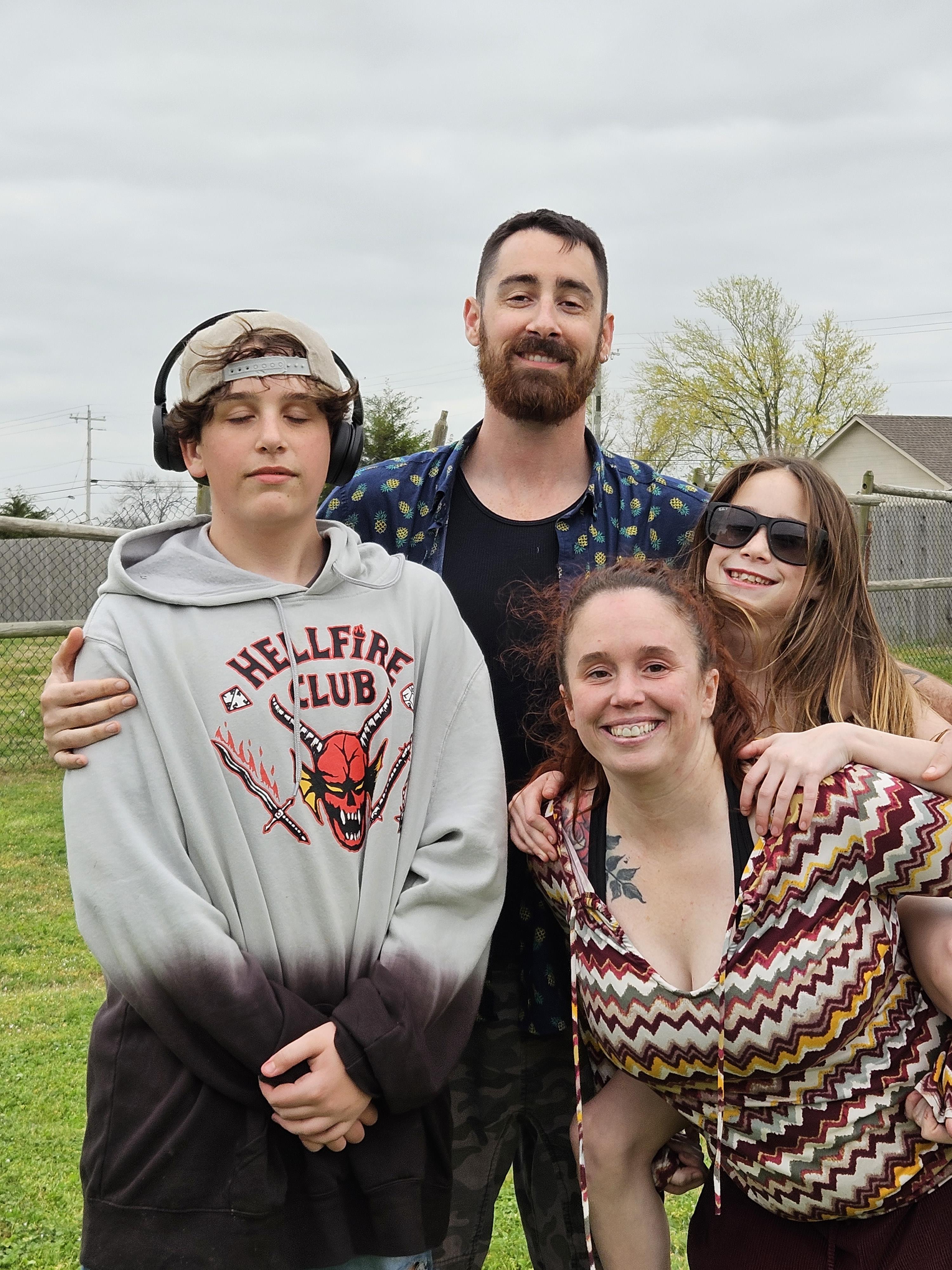 Friends stand together, smiling and dressed casually in a park setting under gray skies.