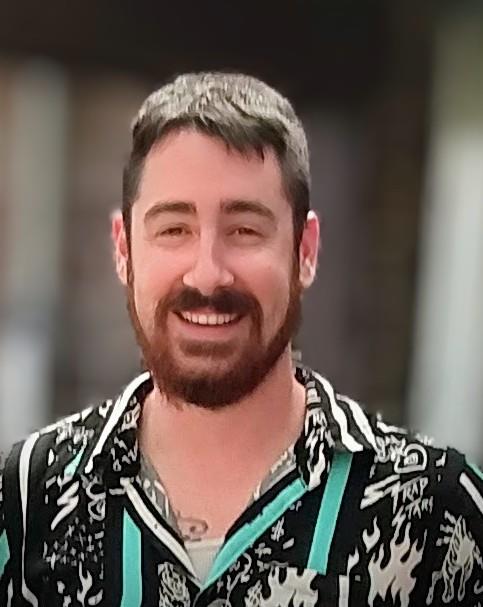 A man with a beard and cheerful expression poses outdoors, dressed in a colorful shirt.