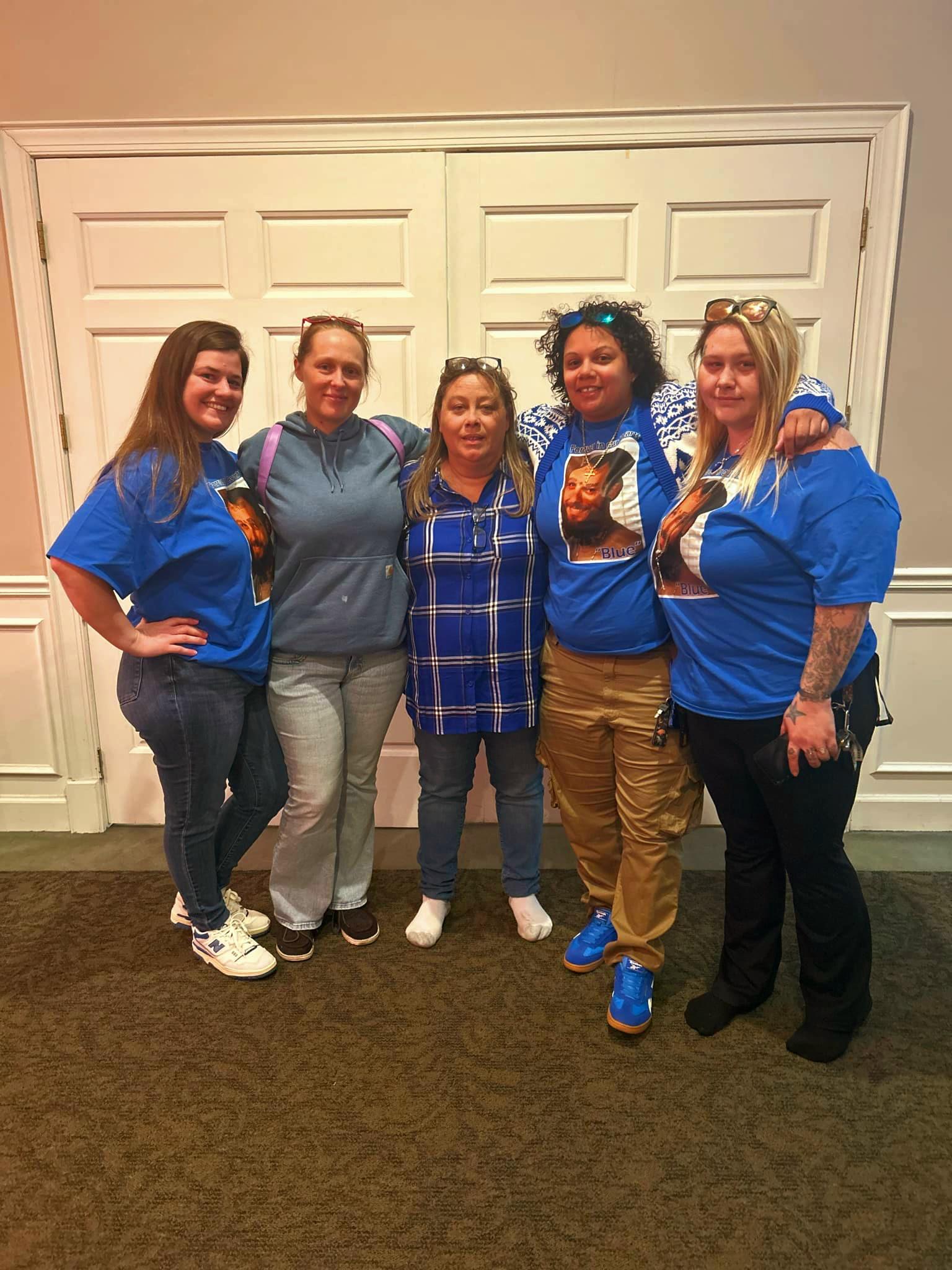 Five women pose together happily, wearing blue shirts with a printed design, at an indoor event.