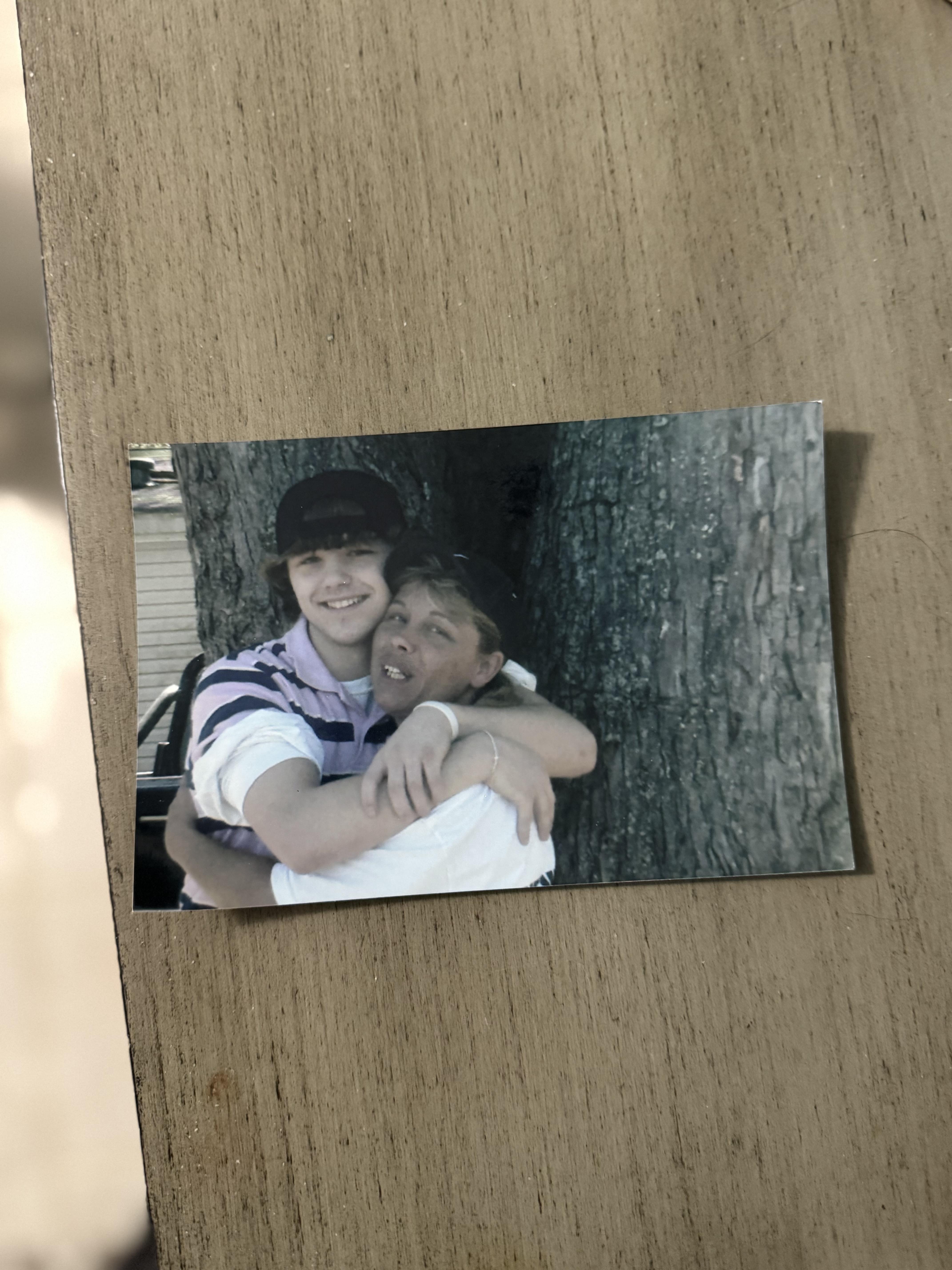 Two friends share a warm embrace outdoors, capturing a joyful memory near a tree.