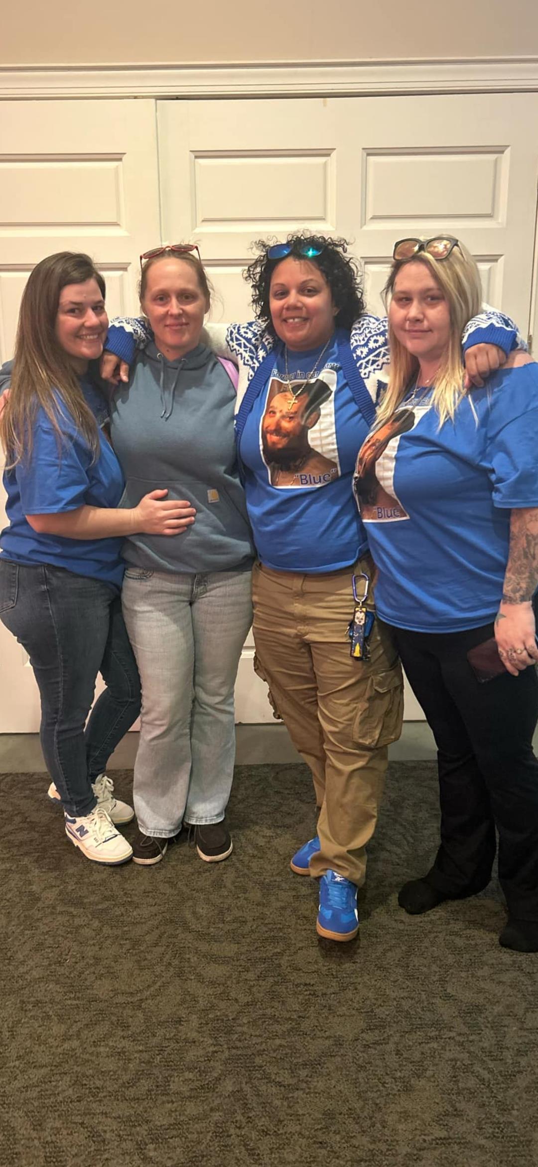 Four friends pose together indoors, wearing matching blue shirts while smiling happily.