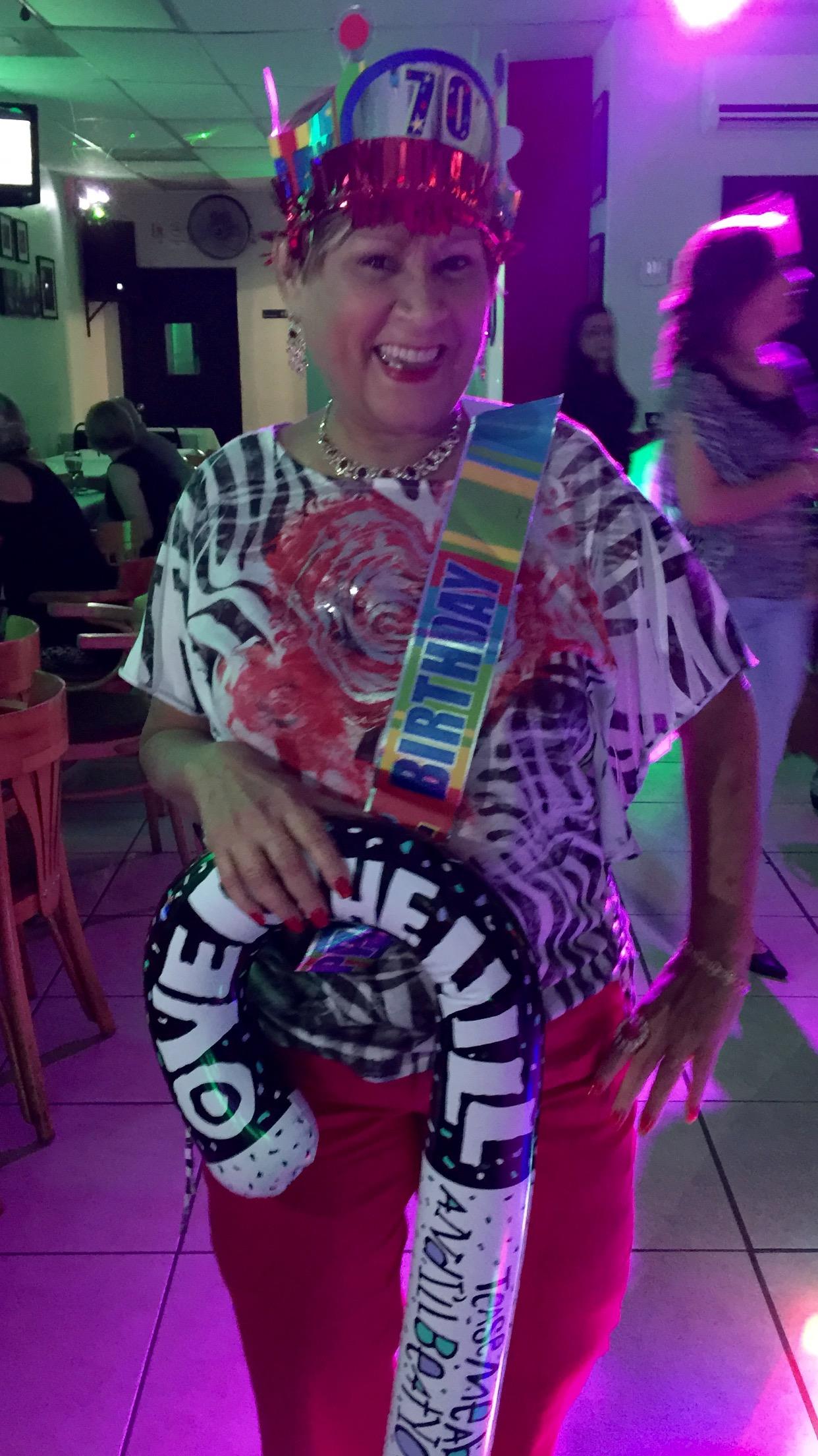 A woman dressed in vibrant attire celebrates her 70th birthday with a big smile and decorations.