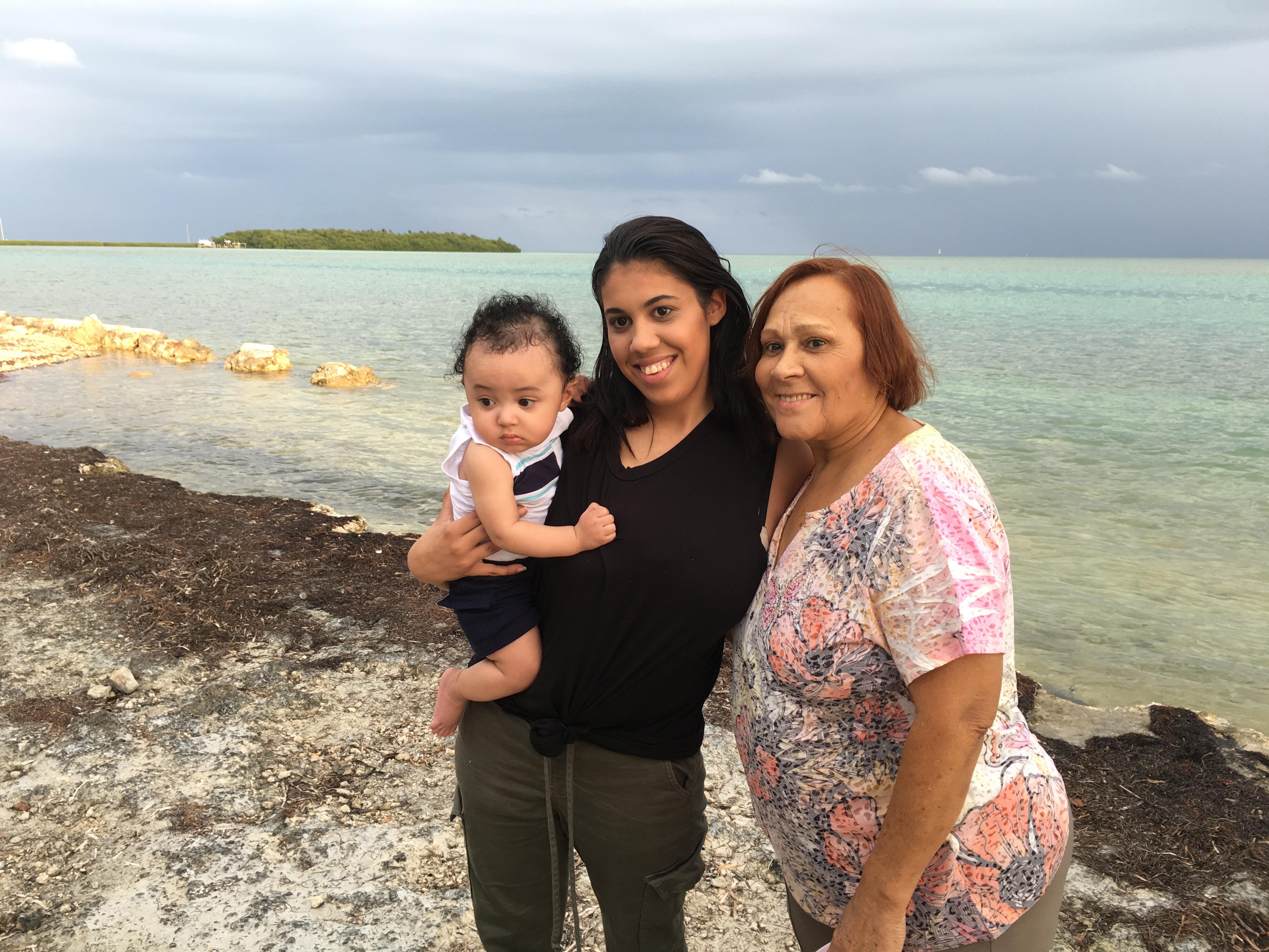 A woman holds a baby while posing with an older lady at the shoreline, showcasing family bonding.