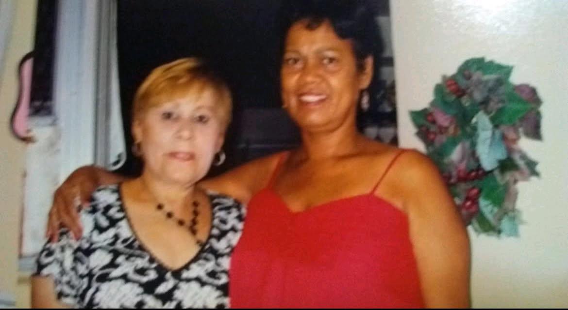 Two women embrace each other while smiling in a welcoming indoor setting decorated with plants.