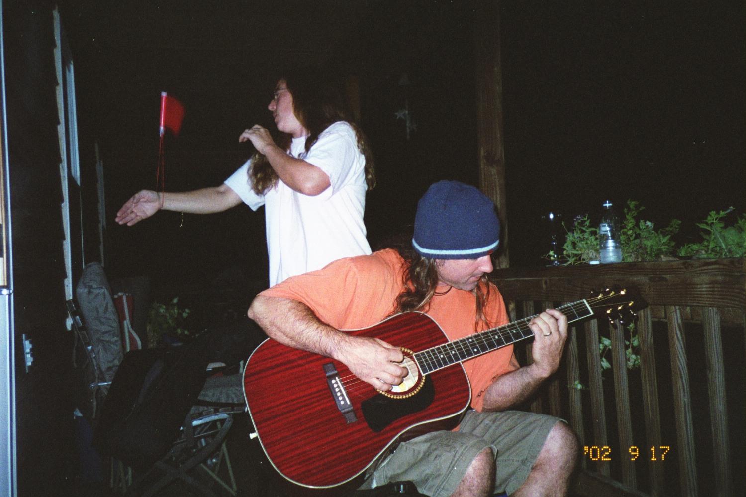 A man plays guitar while a friend interacts nearby, enjoying a relaxed evening outdoors.